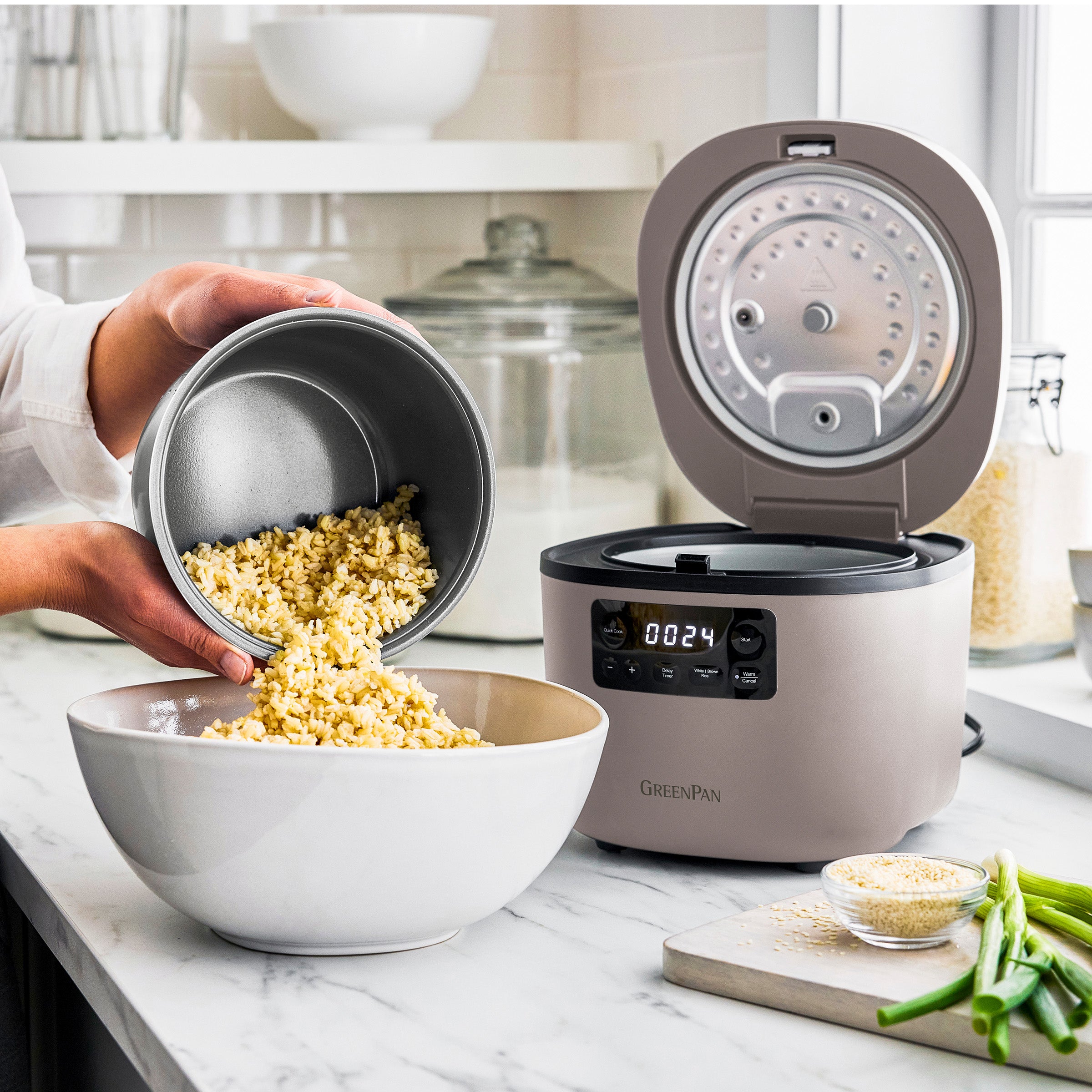 Person transferring cooked rice from a small container into a bowl next to a rice cooker on a kitchen counter.