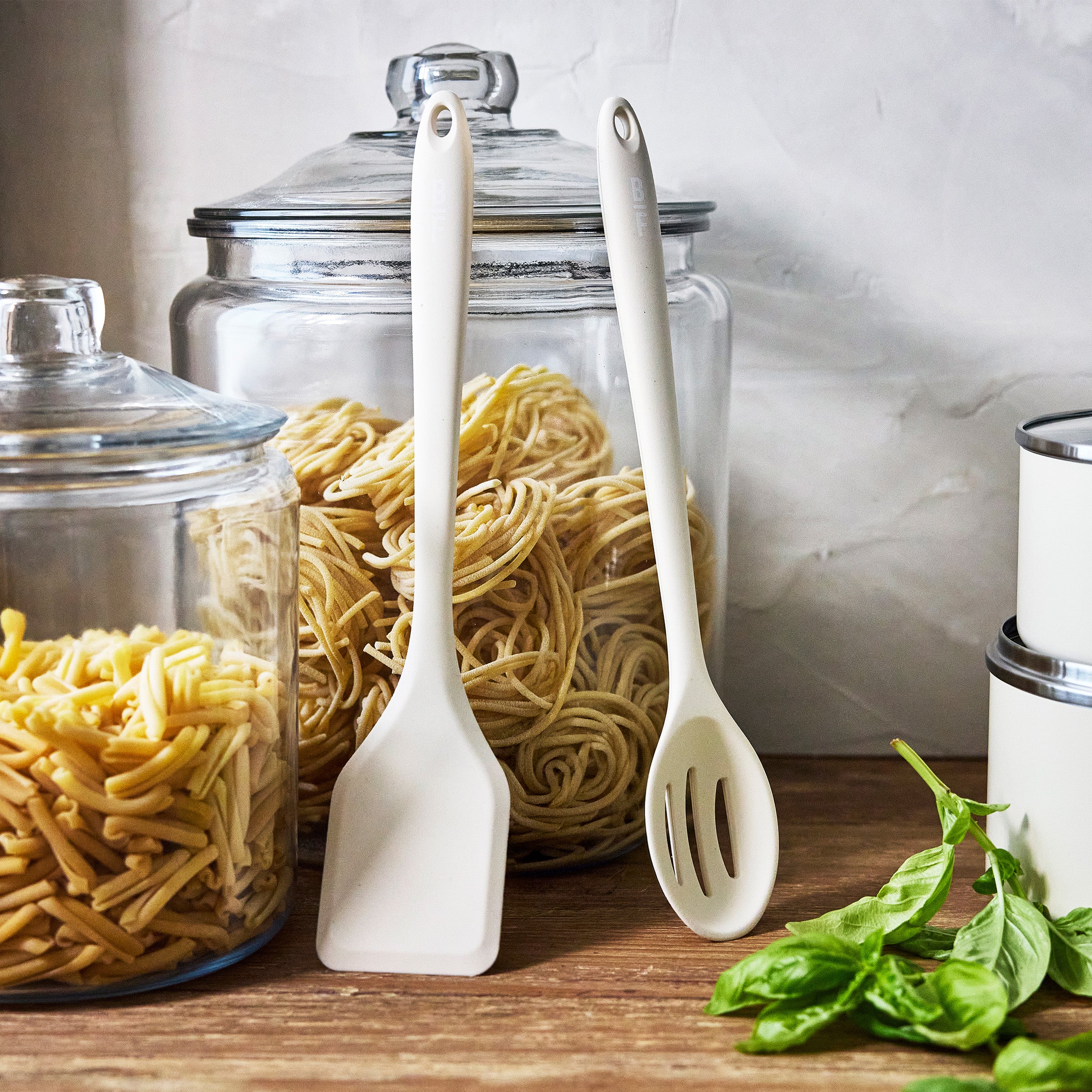Cream cookware set with metal handles on wooden surface, with jars of dry pasta and kitchen utensils in the background.