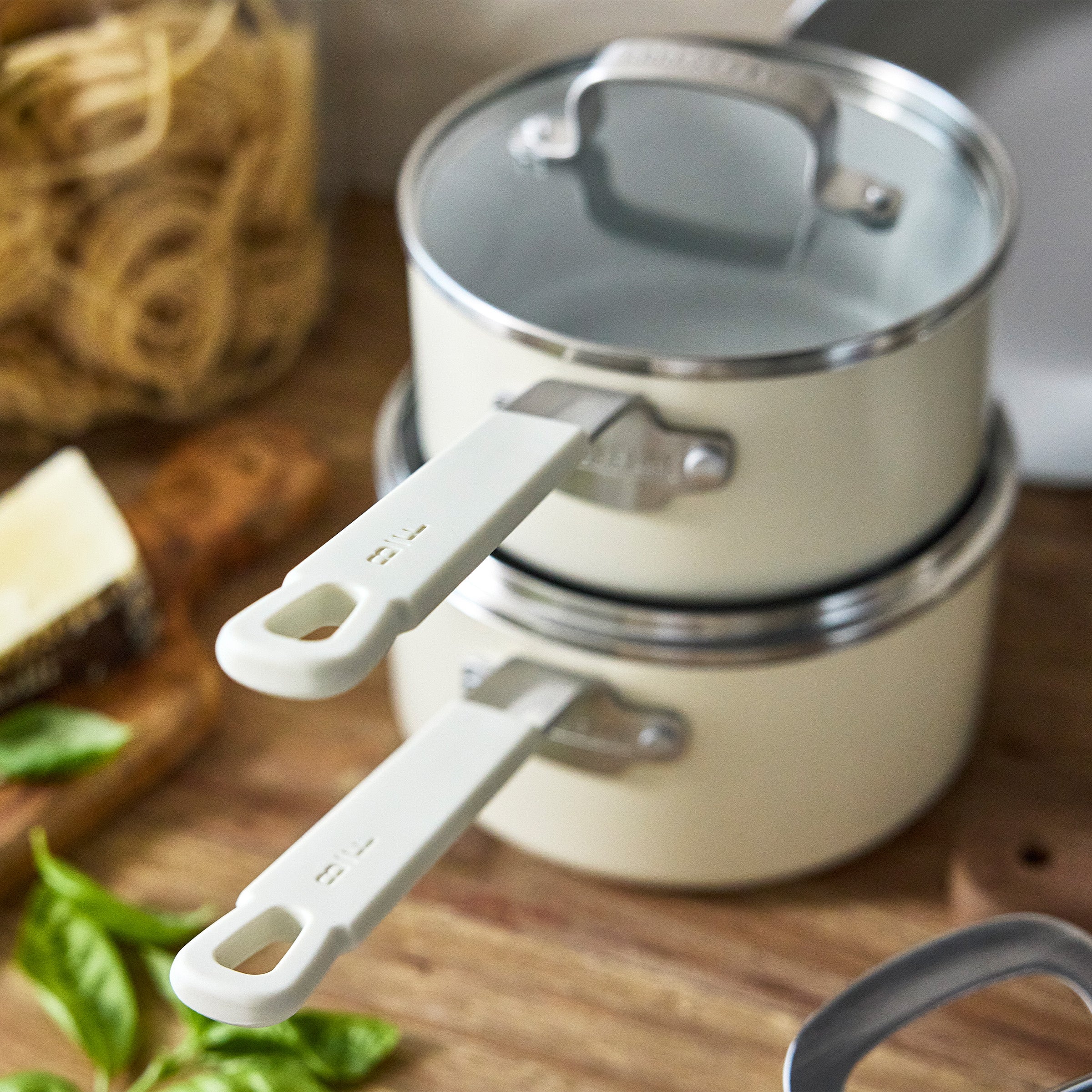 Two cream-colored pans stacked on a wooden counter with pasta, fresh basil, and cheese nearby.