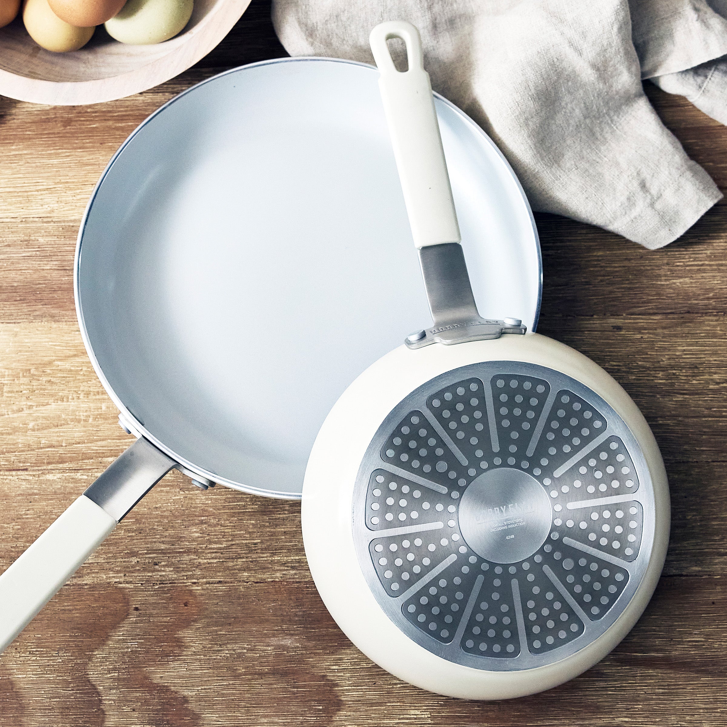 Two cream-colored frying pans, one facing up and one down, on wood with a beige cloth and a bowl of eggs nearby.