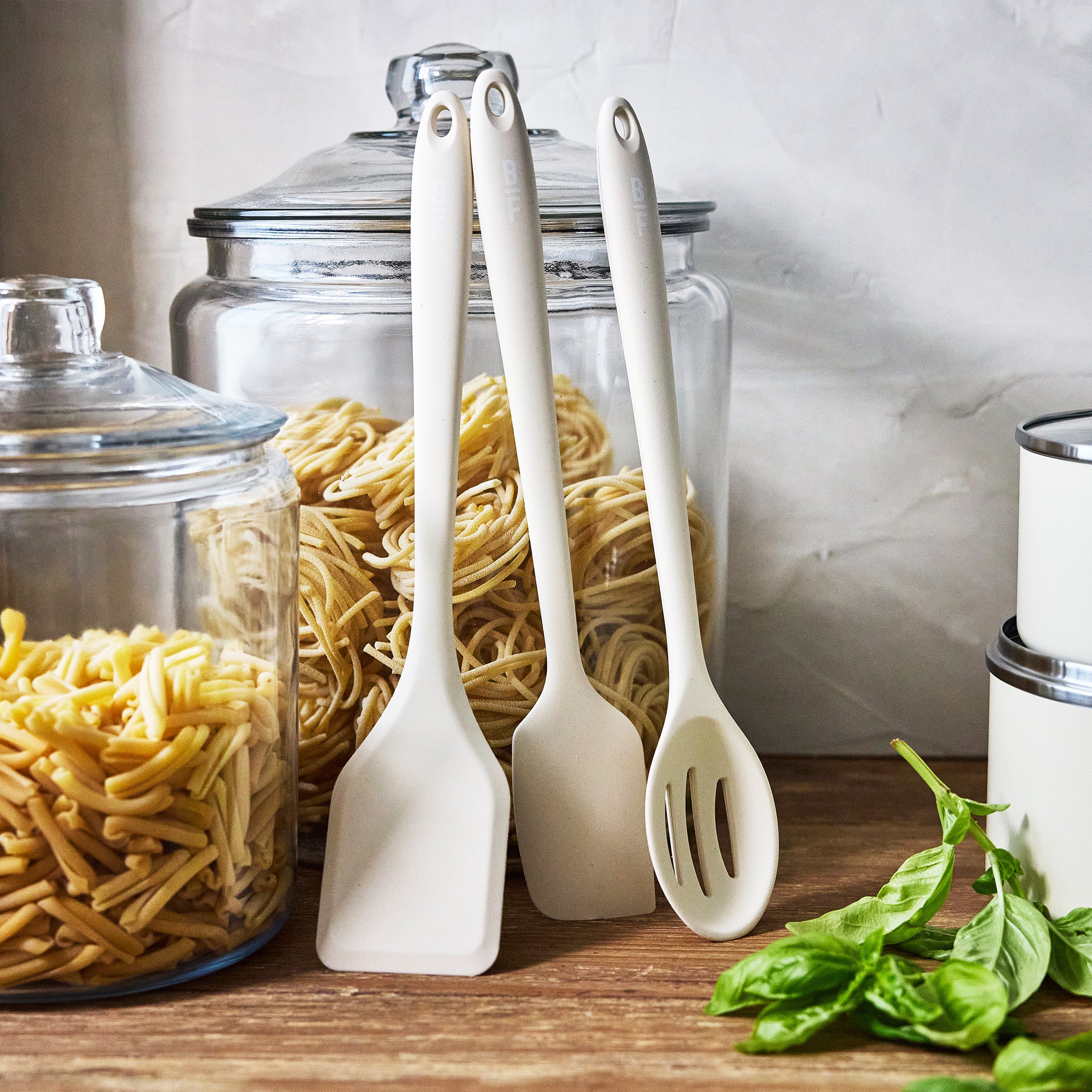 Three cream kitchen utensils next to clear pasta jars and a basil plant on a kitchen counter.