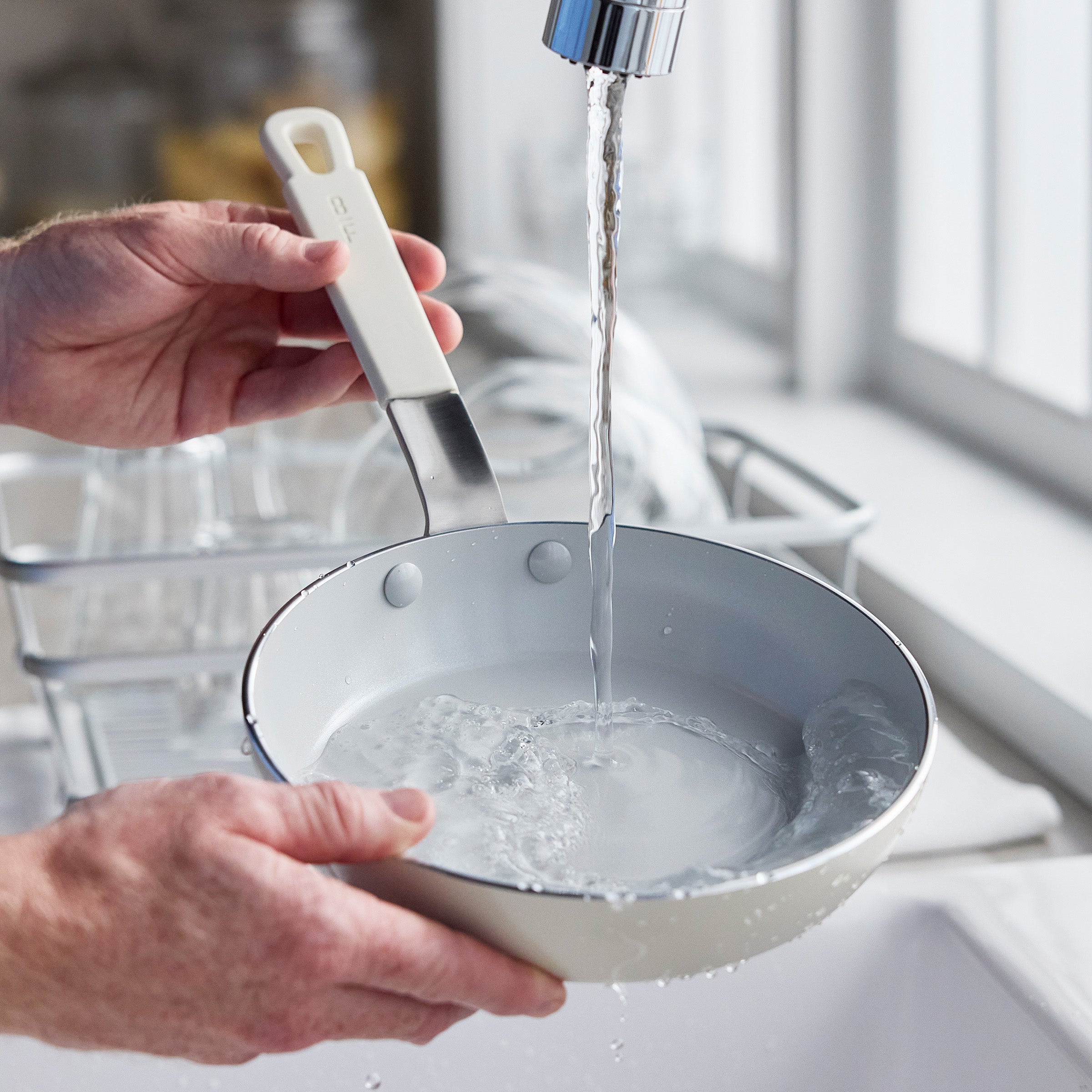 Cream-colored frying pan rinsed under water at a kitchen sink, dish rack visible nearby.