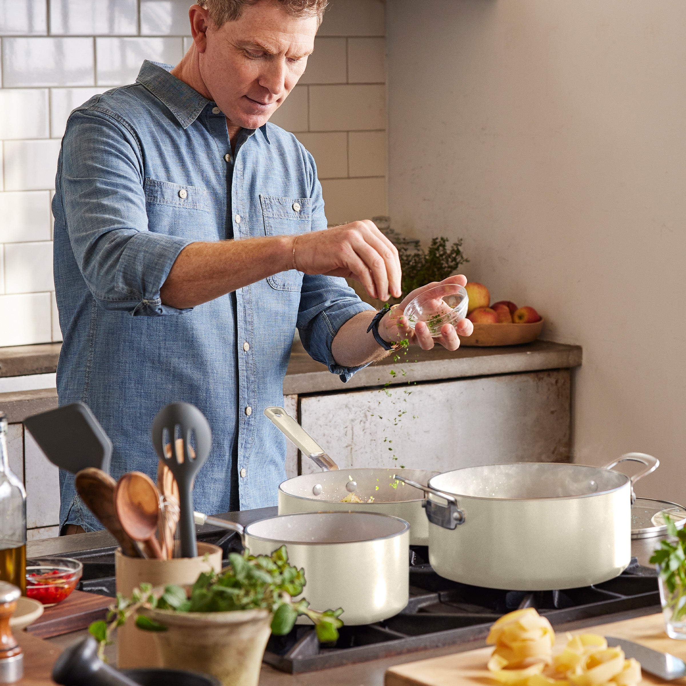 Man in a denim shirt adds herbs to a GreenPan ceramic nonstick pot with fresh ingredients on the stove.