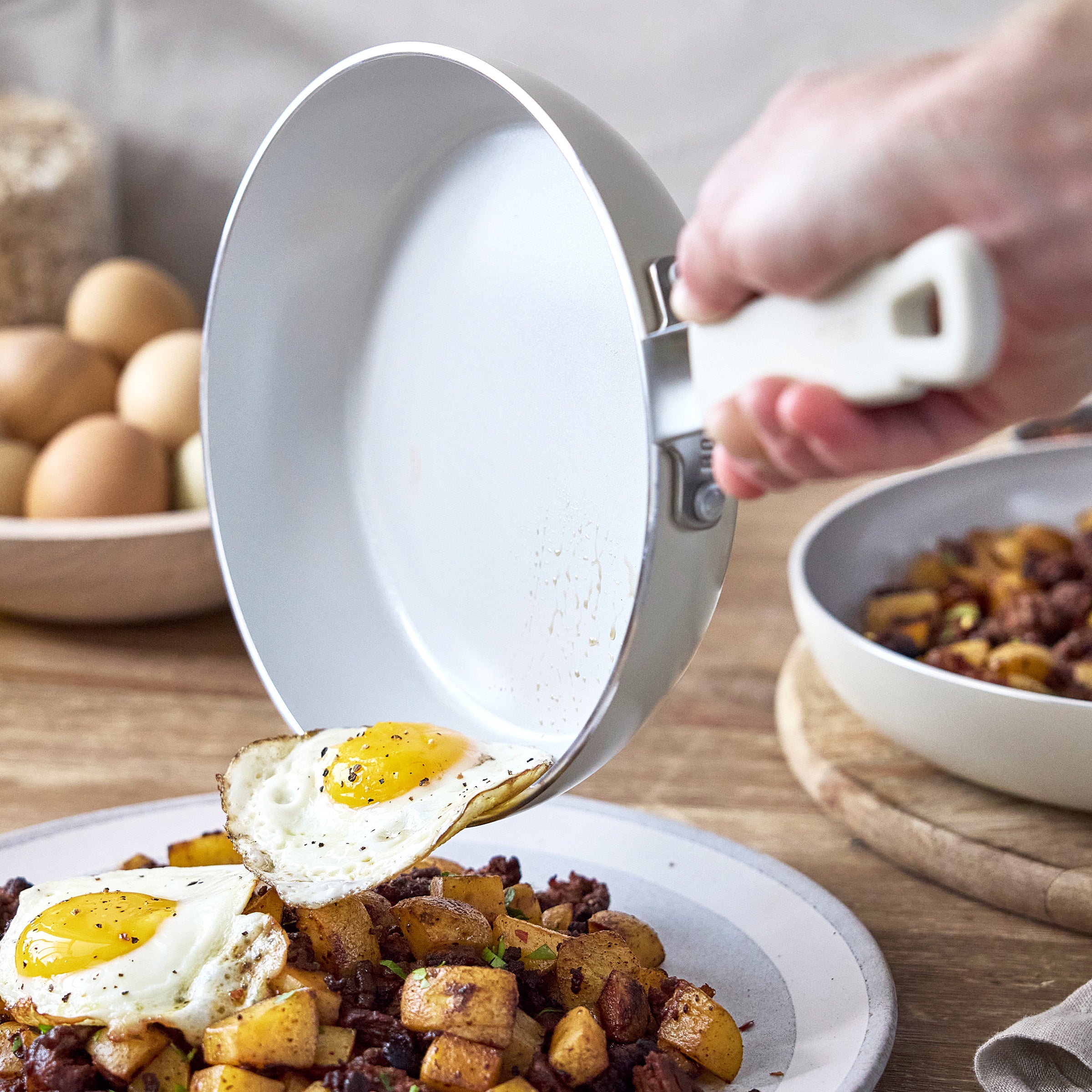 Hand sliding two fried eggs from a cream-colored nonstick fry pan onto a plate with potatoes and meat, bowls in the background.