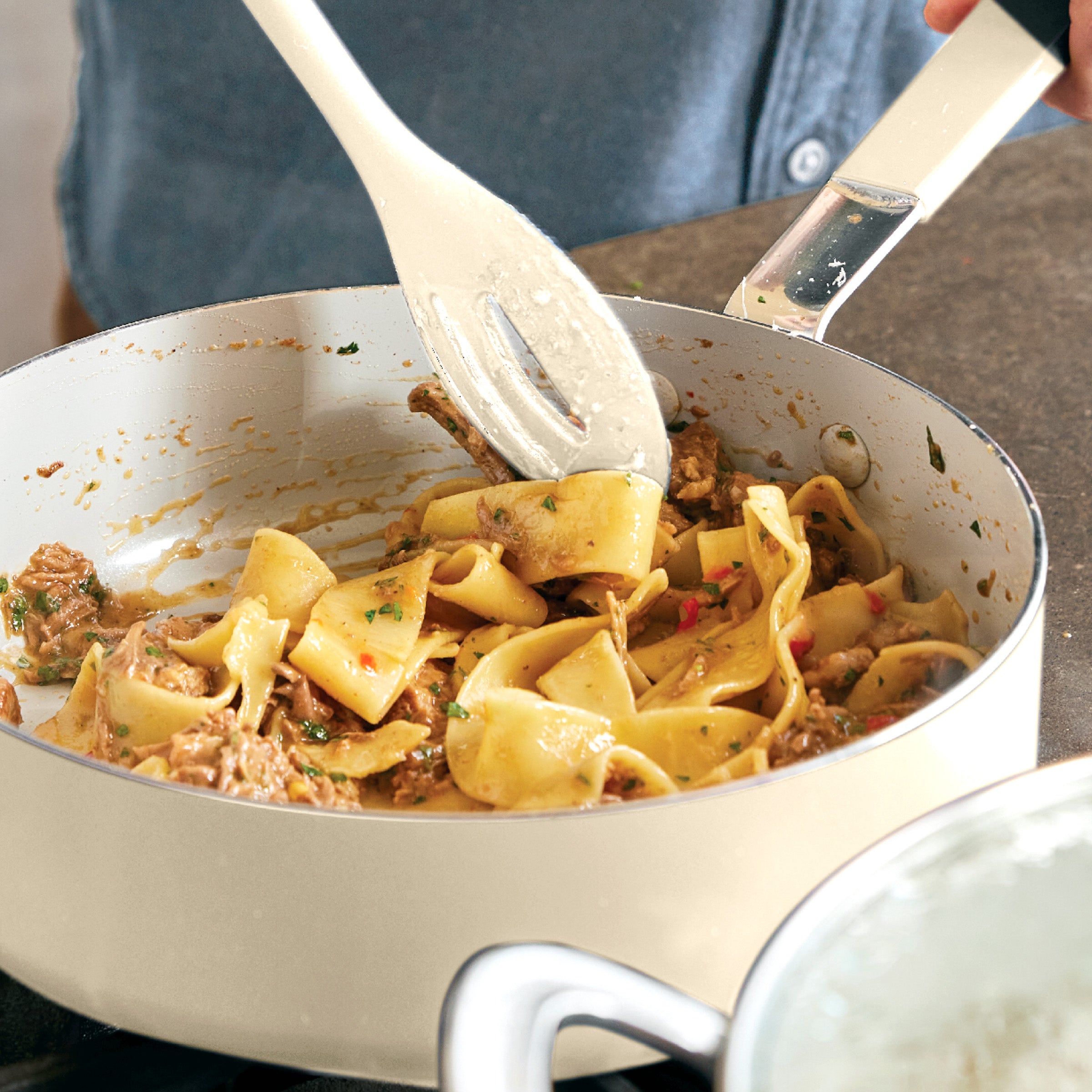 Person in blue shirt stirs pasta in a cream Bobby Flay by GreenPan pot from a visible 16-piece cookware set.