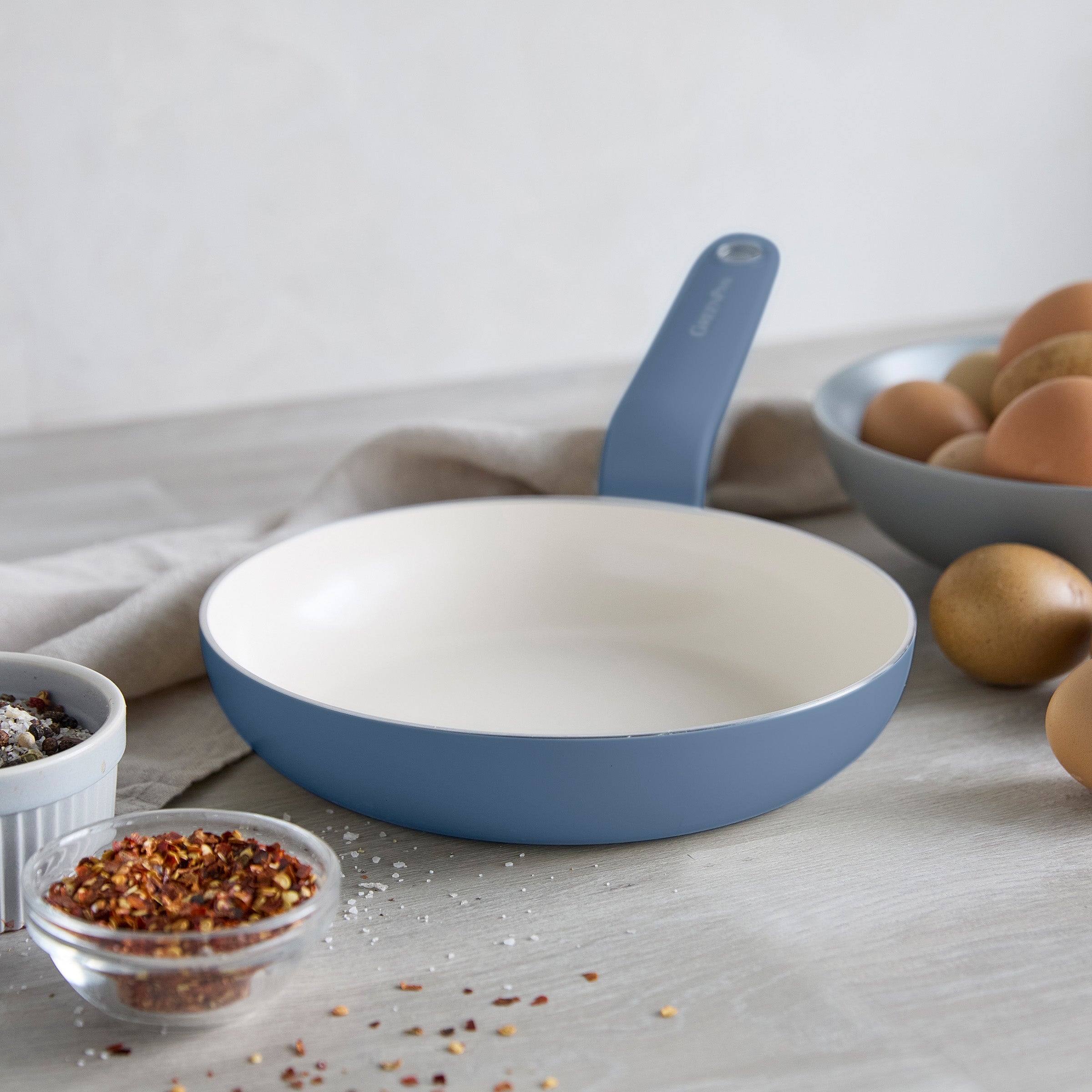 Blue ceramic nonstick 8-inch frypan on a counter with eggs, spices, and a bowl of ingredients nearby.