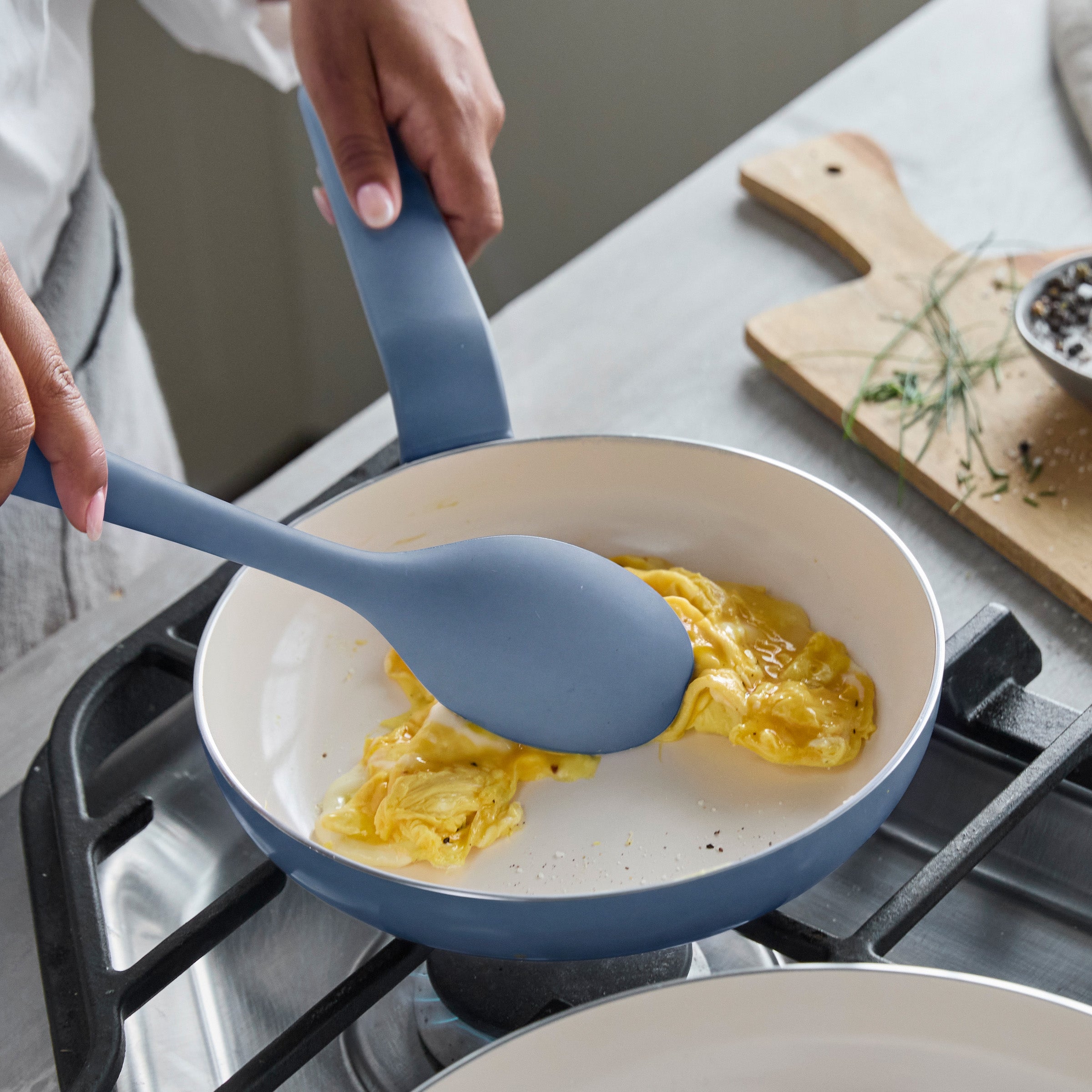 Scrambled eggs in a blue ceramic nonstick 8-inch frypan, with a blue spatula and fresh herbs beside it.