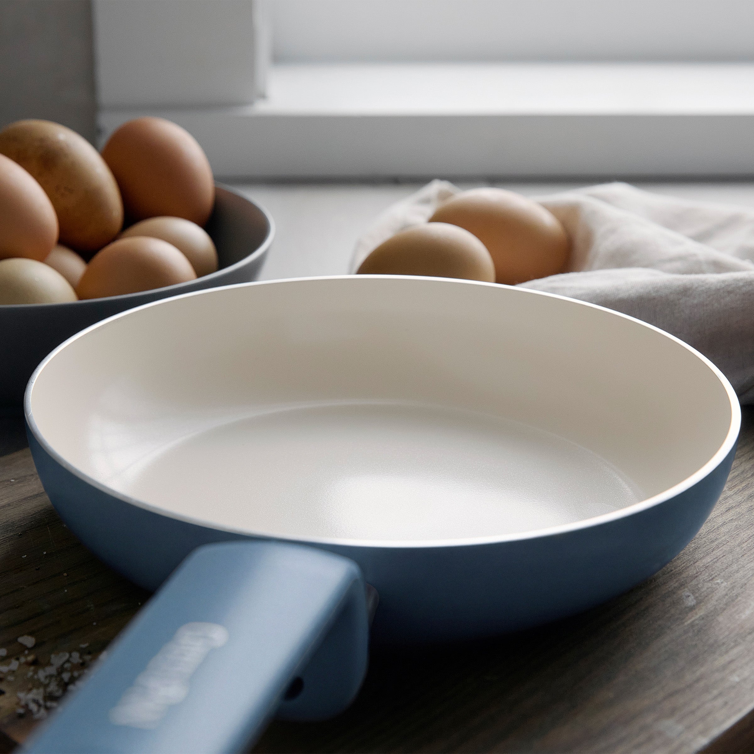 Blue ceramic 8-inch frypan on a counter with eggs and a cloth, sunlight streaming through a nearby window.