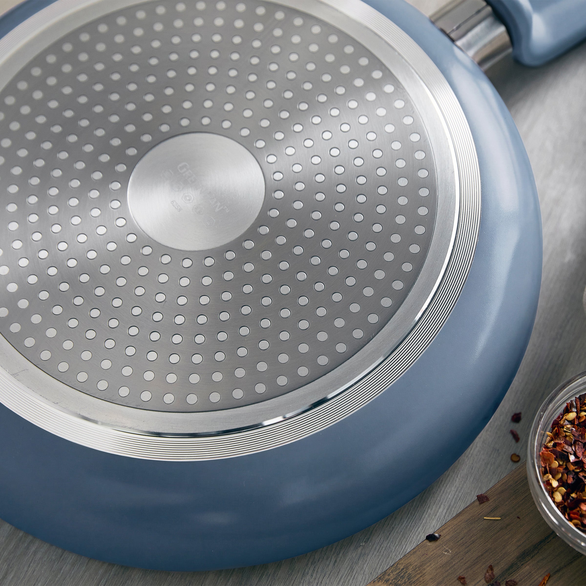 Blue ceramic 8-inch frypan with textured metal base, placed on wood beside a small bowl of red chili flakes.