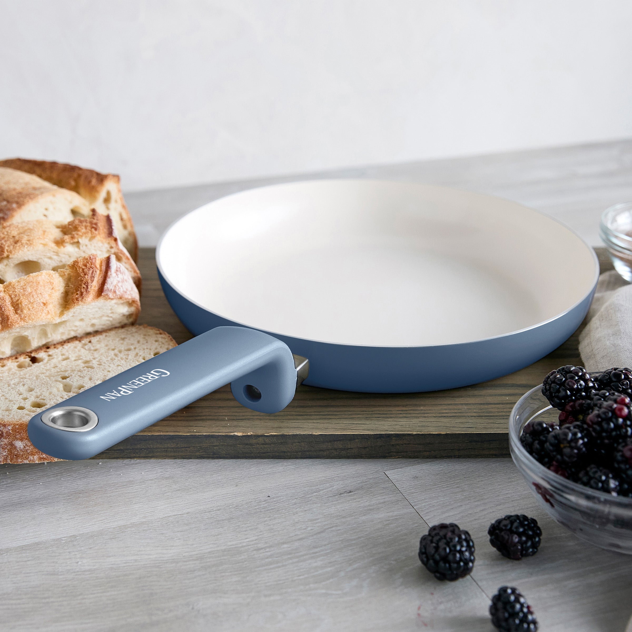 Blue ceramic frying pan with bread and blackberries on a wooden board, placed on a light-colored countertop.