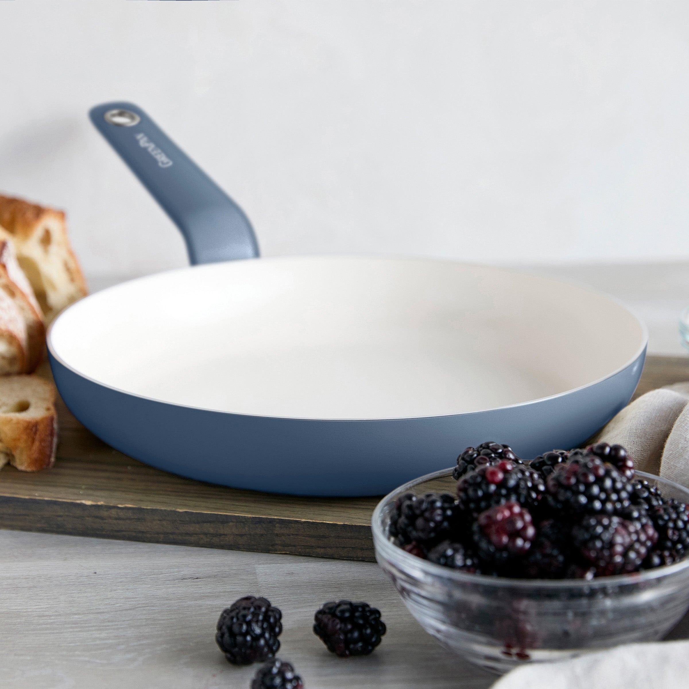Blue ceramic nonstick frypan on a wooden board with blackberries and sliced bread nearby.
