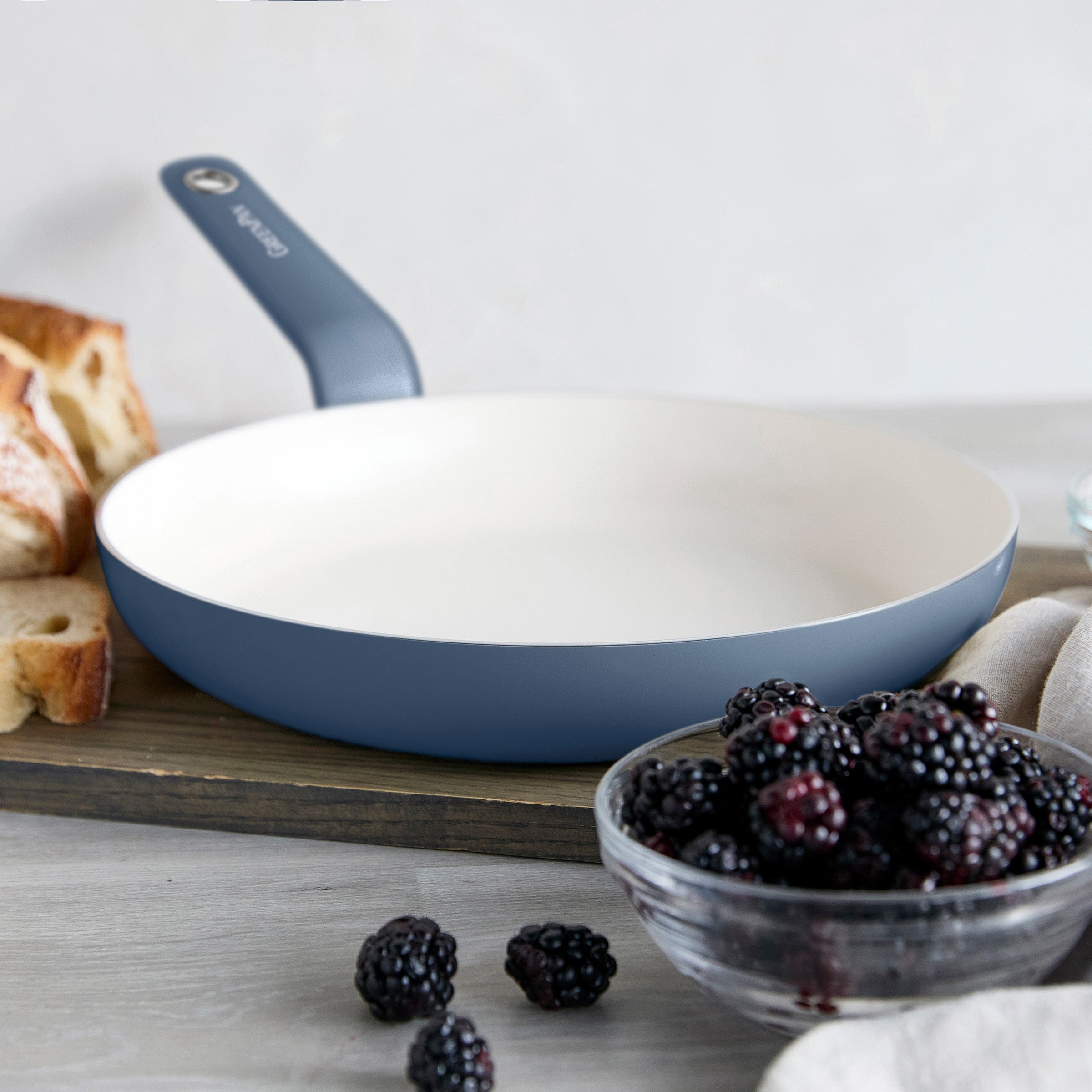 Blue ceramic 12-inch frypan on a wooden board with blackberries and bread slices nearby.