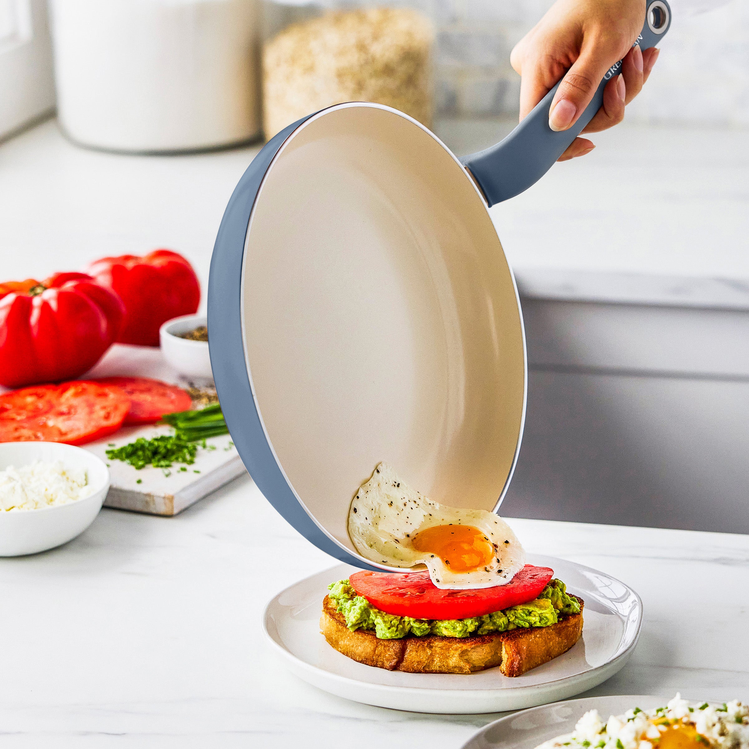 Fried egg being slid from a blue ceramic nonstick frying pan onto avocado toast by a hand in a bright kitchen.