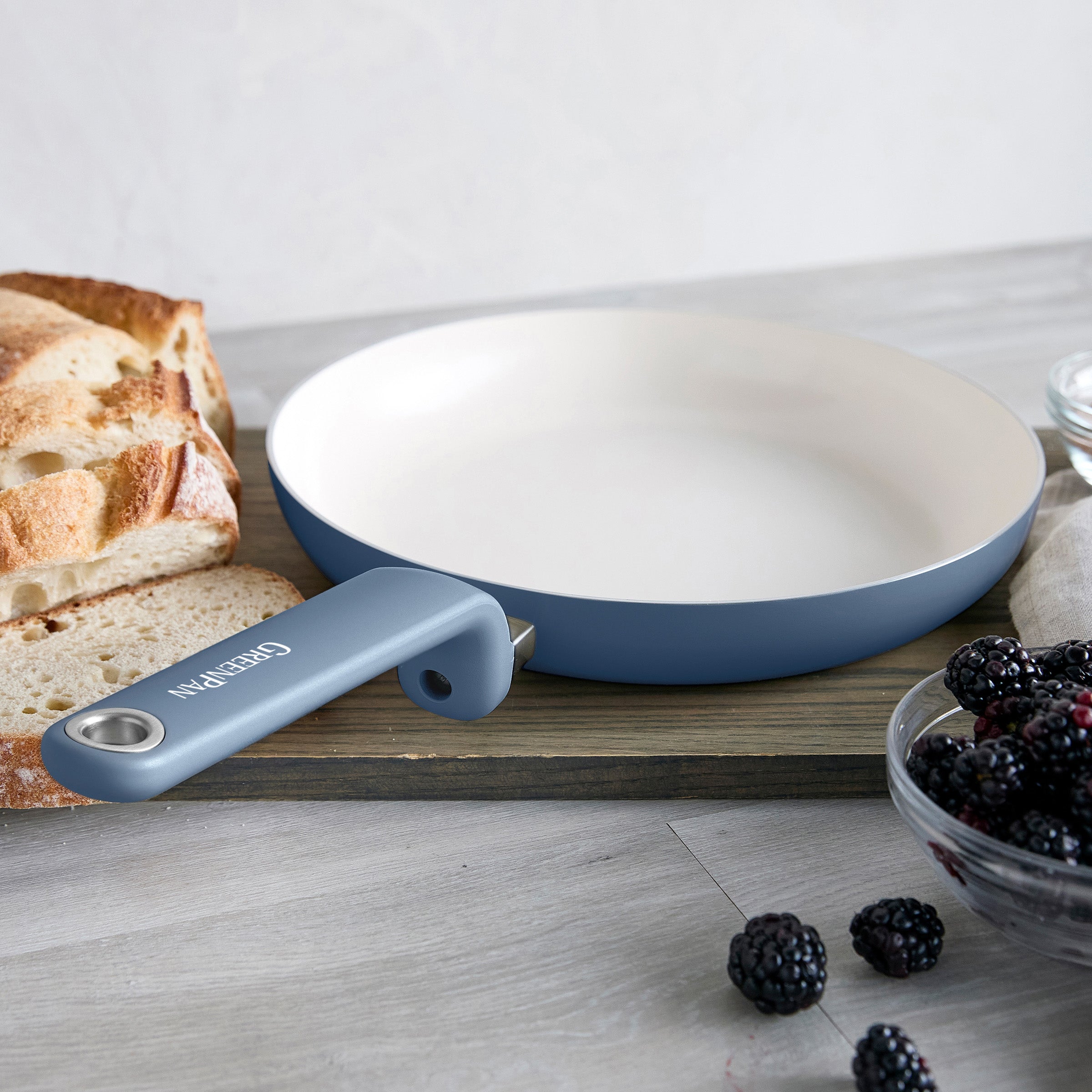 Blue ceramic frypan with bread and blackberries on a wooden board and light surface.