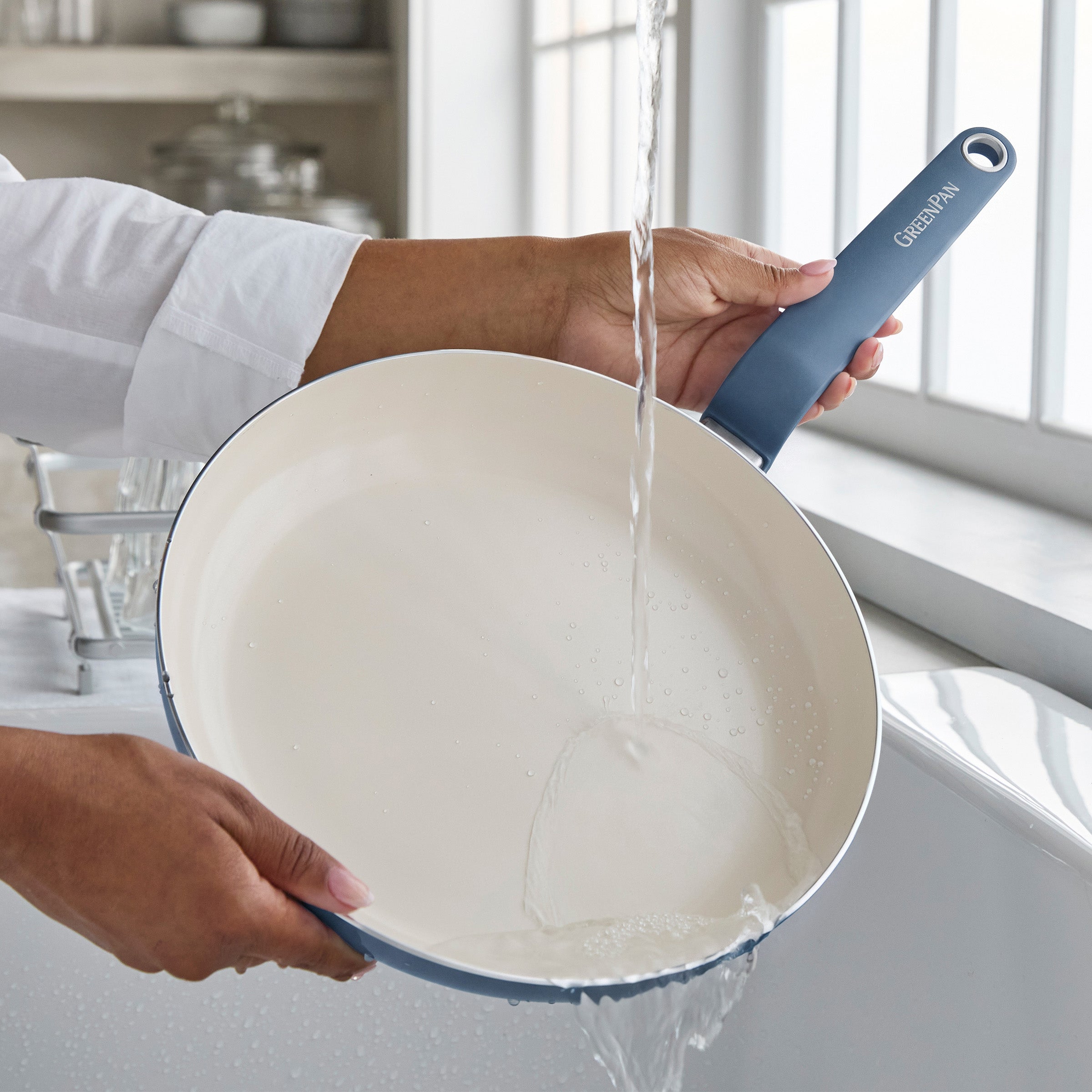 Person rinsing a blue ceramic 12-inch frypan under running water in a bright kitchen near a window.