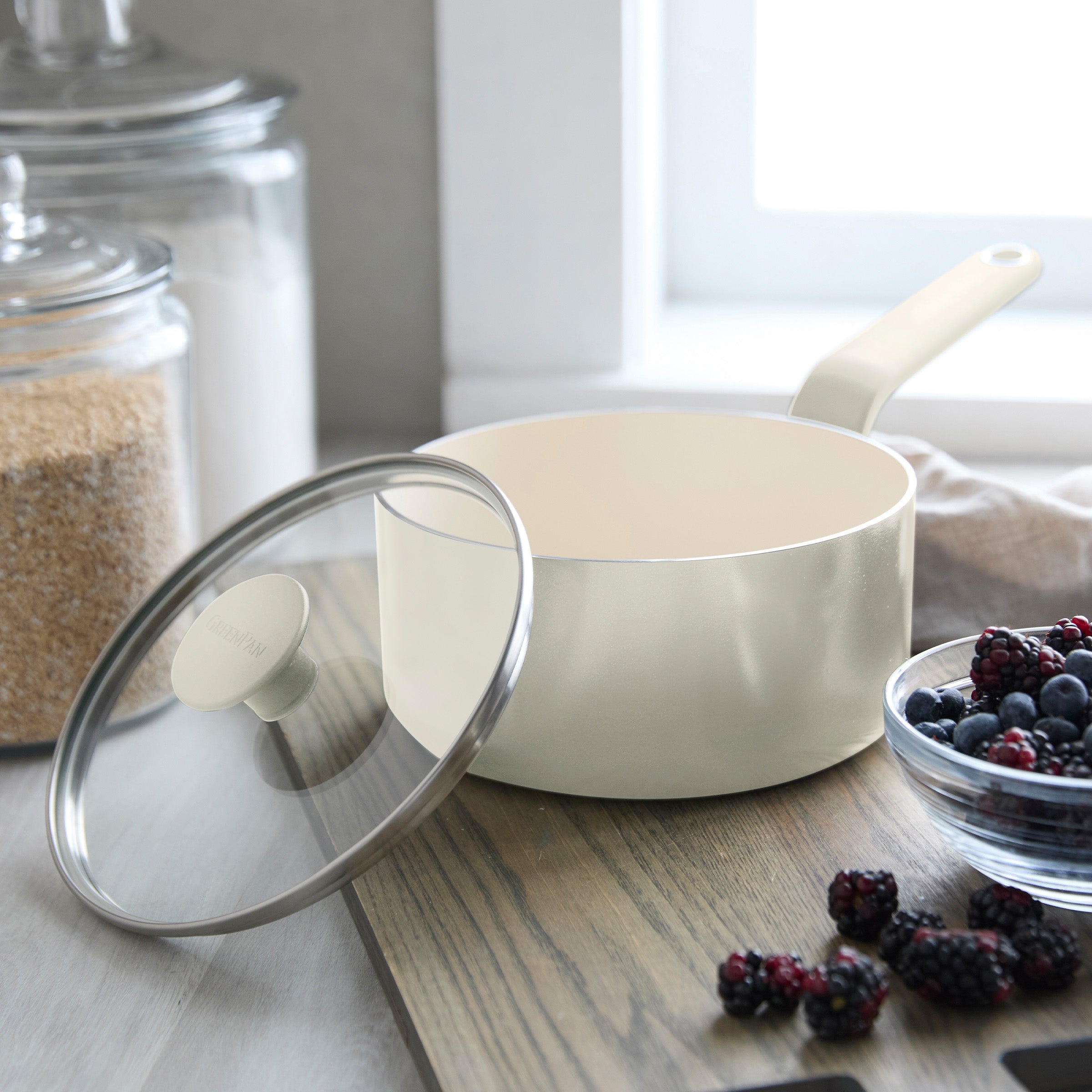 Cream ceramic 2-quart saucepan with lid on wood counter, next to berries and glass jars filled with sugar.