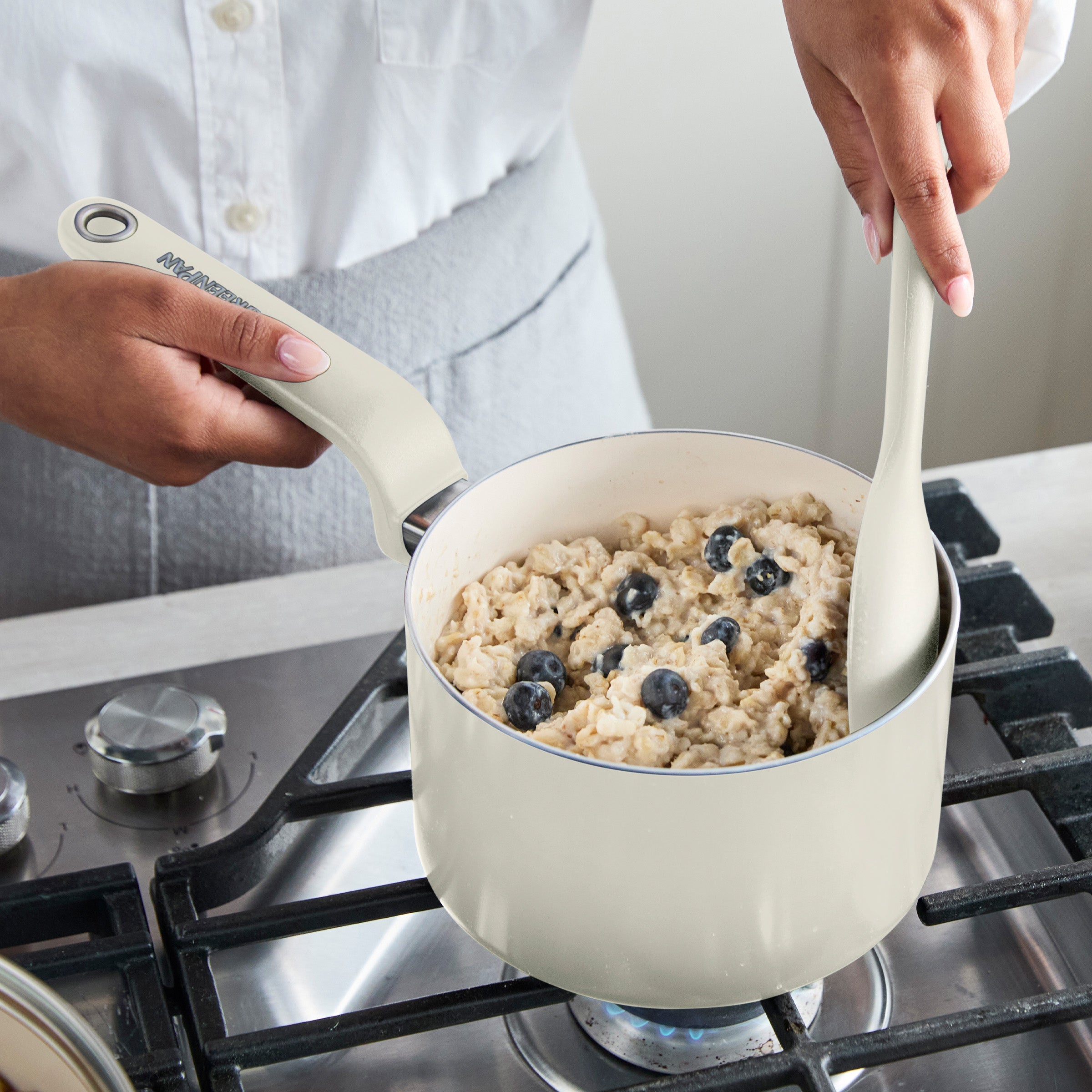 Cream ceramic saucepan with lid on a stove, blueberry oatmeal being stirred inside.