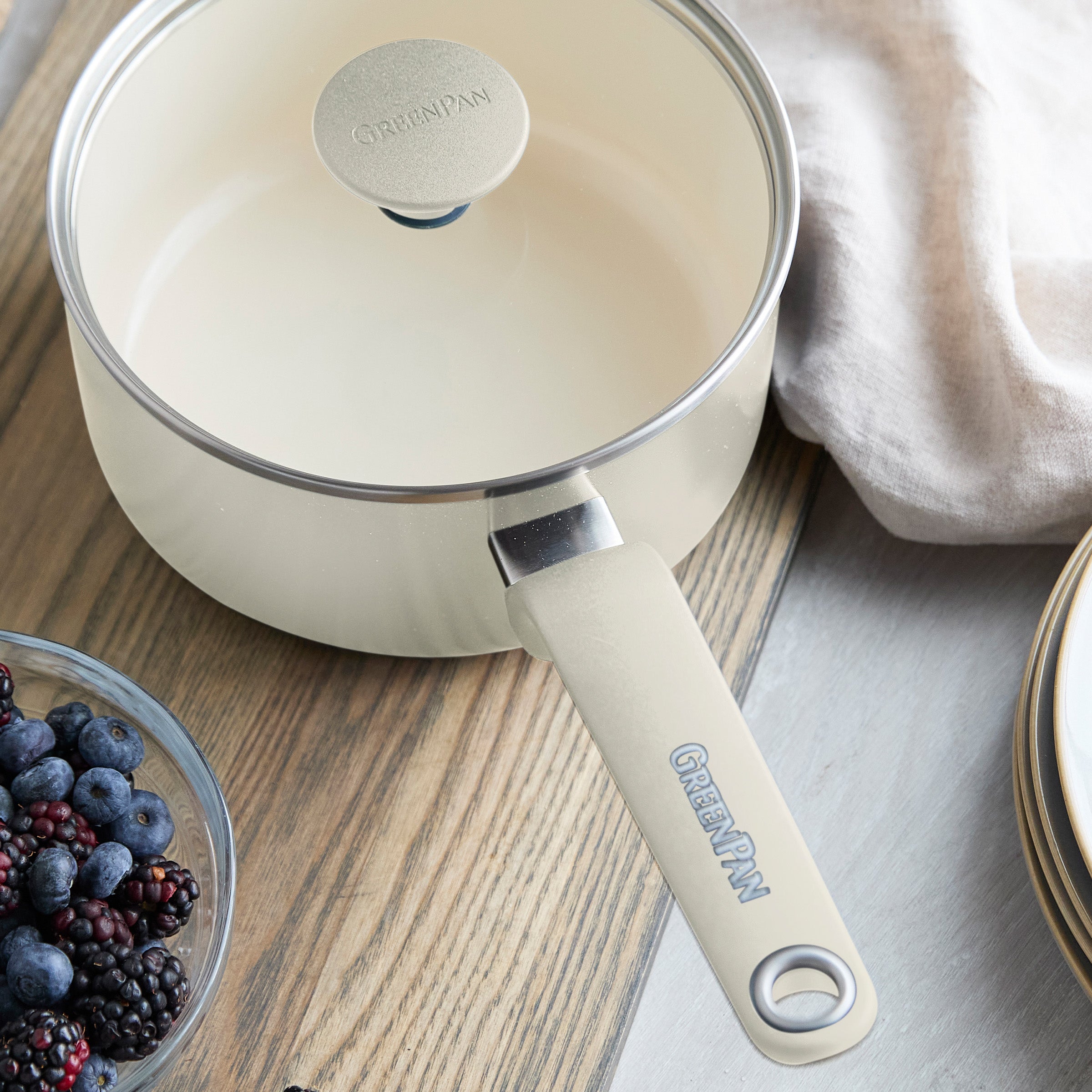 Cream ceramic 2-quart saucepan with lid on wooden table, shown beside berries, plates, and a beige napkin.
