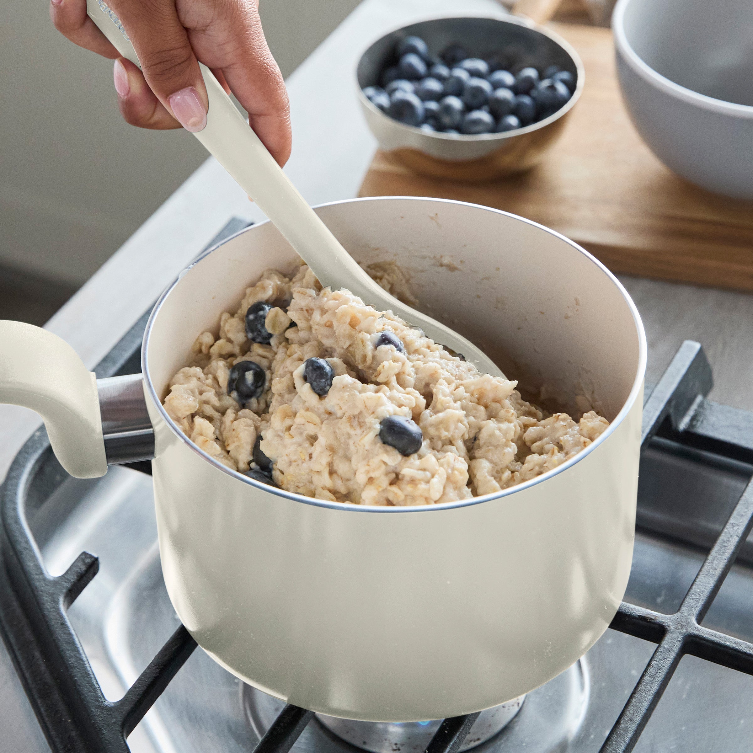 Hand stirring oatmeal with blueberries in a gray ceramic saucepan; fresh blueberries beside the pan on a white surface.