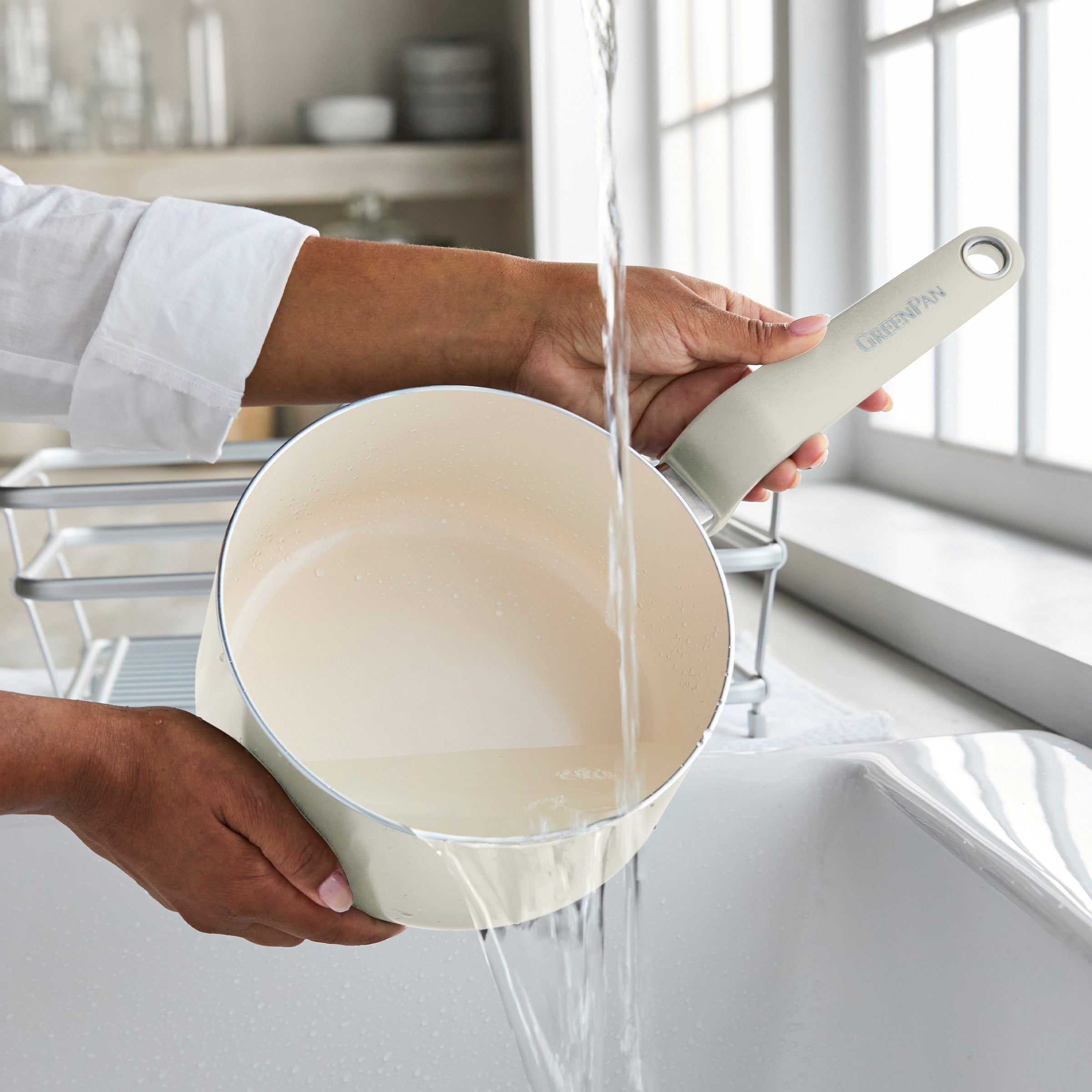 A cream ceramic nonstick saucepan with lid being washed under running water by a kitchen sink.