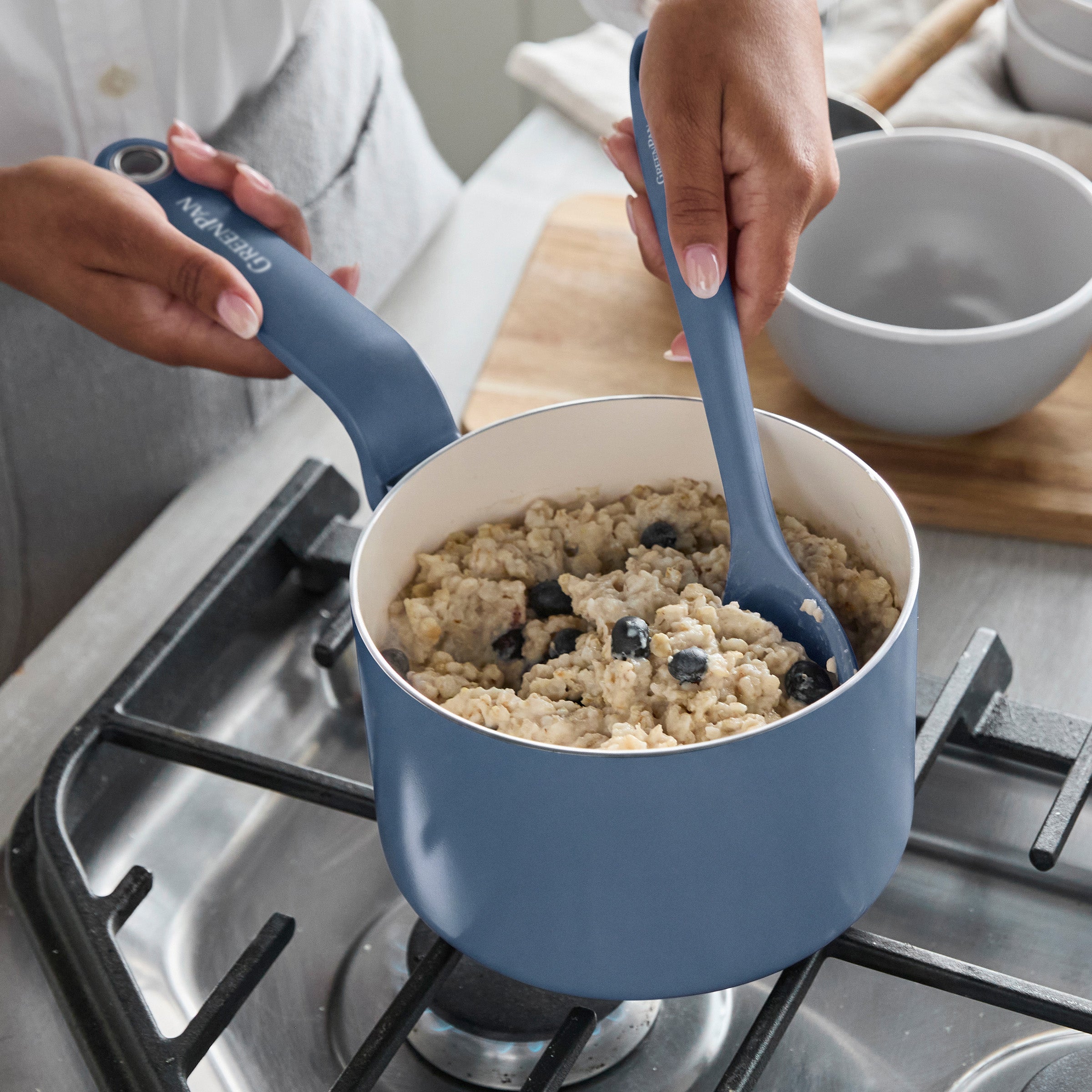 Blue ceramic nonstick saucepan with lid on stovetop, oatmeal being stirred inside.