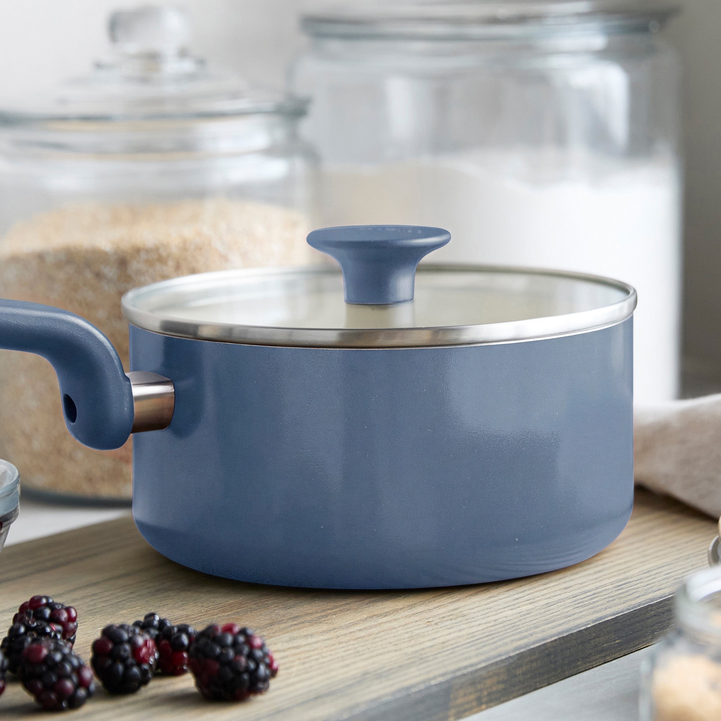 Blue ceramic 2-quart saucepan with lid on a board, surrounded by blackberries and glass jars.