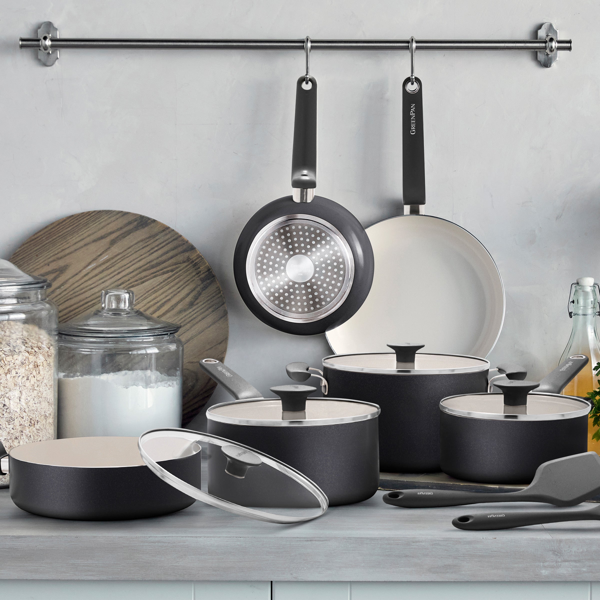 Black 12-piece ceramic nonstick cookware set with glass lids, plus two matching pans hanging on a metal rack above.