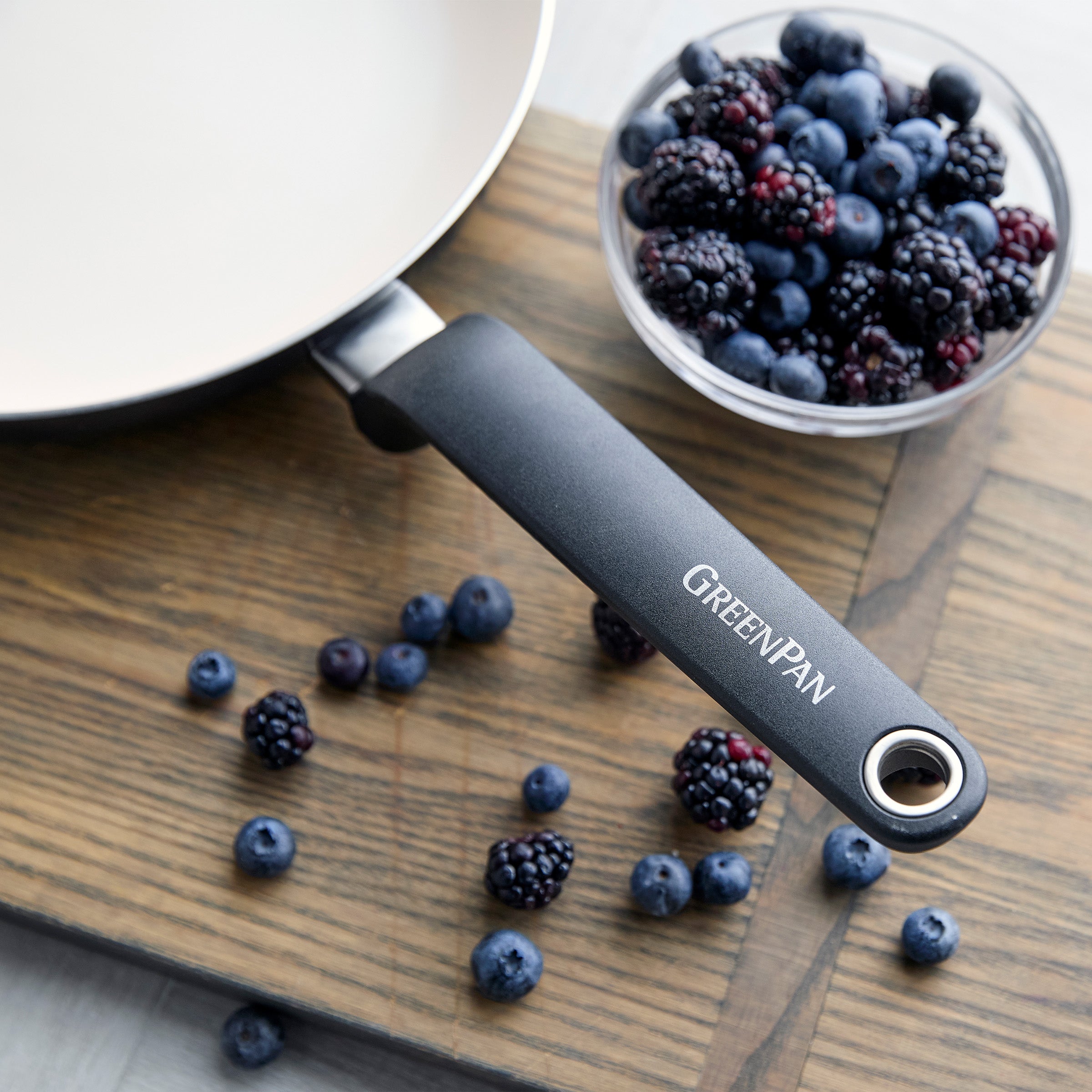 Rio 12-piece ceramic nonstick cookware skillet beside a bowl of mixed berries on a wooden surface, with berries scattered nearby.
