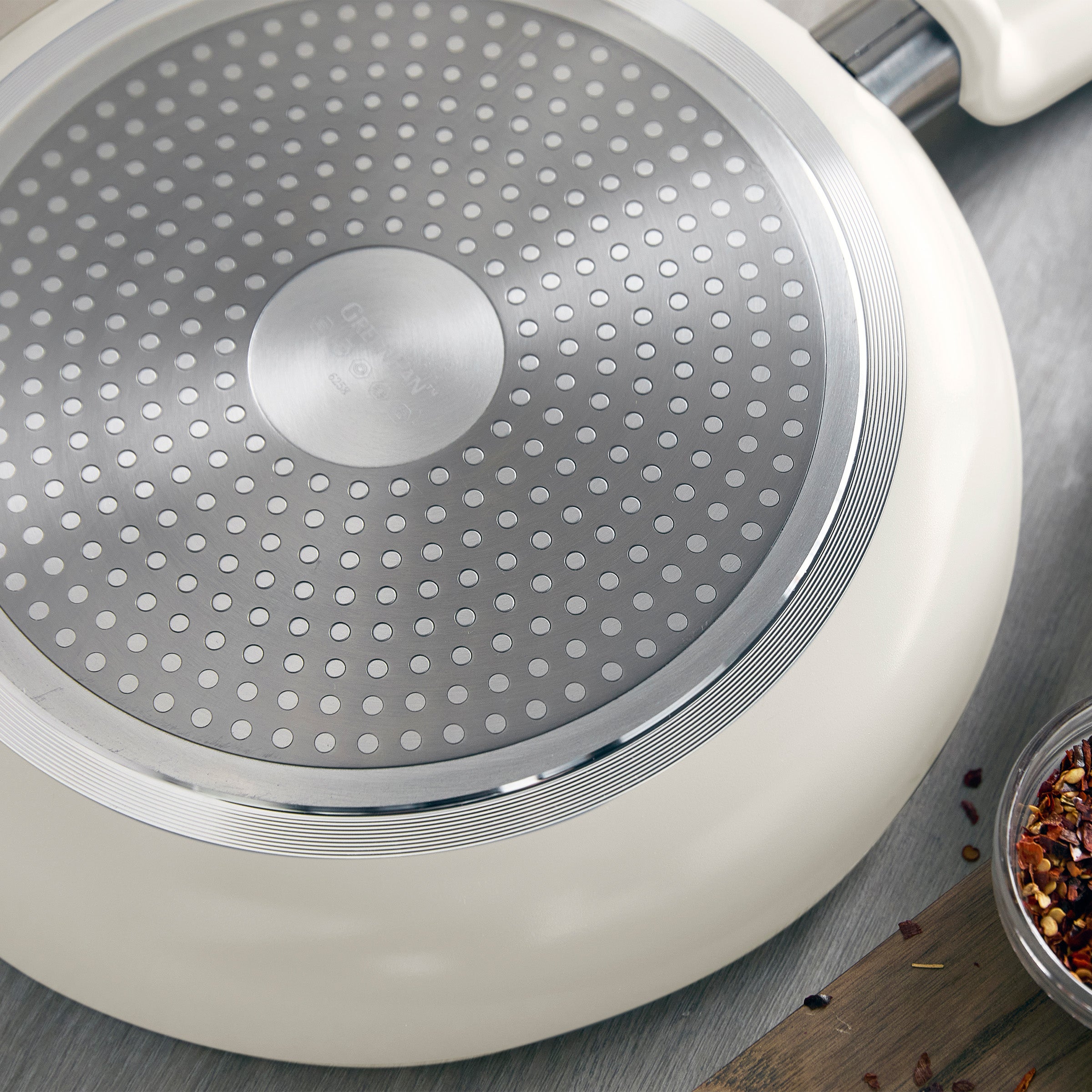 Bottom of a ceramic nonstick frying pan with metal plate and dotted pattern, beside scattered dried spices.