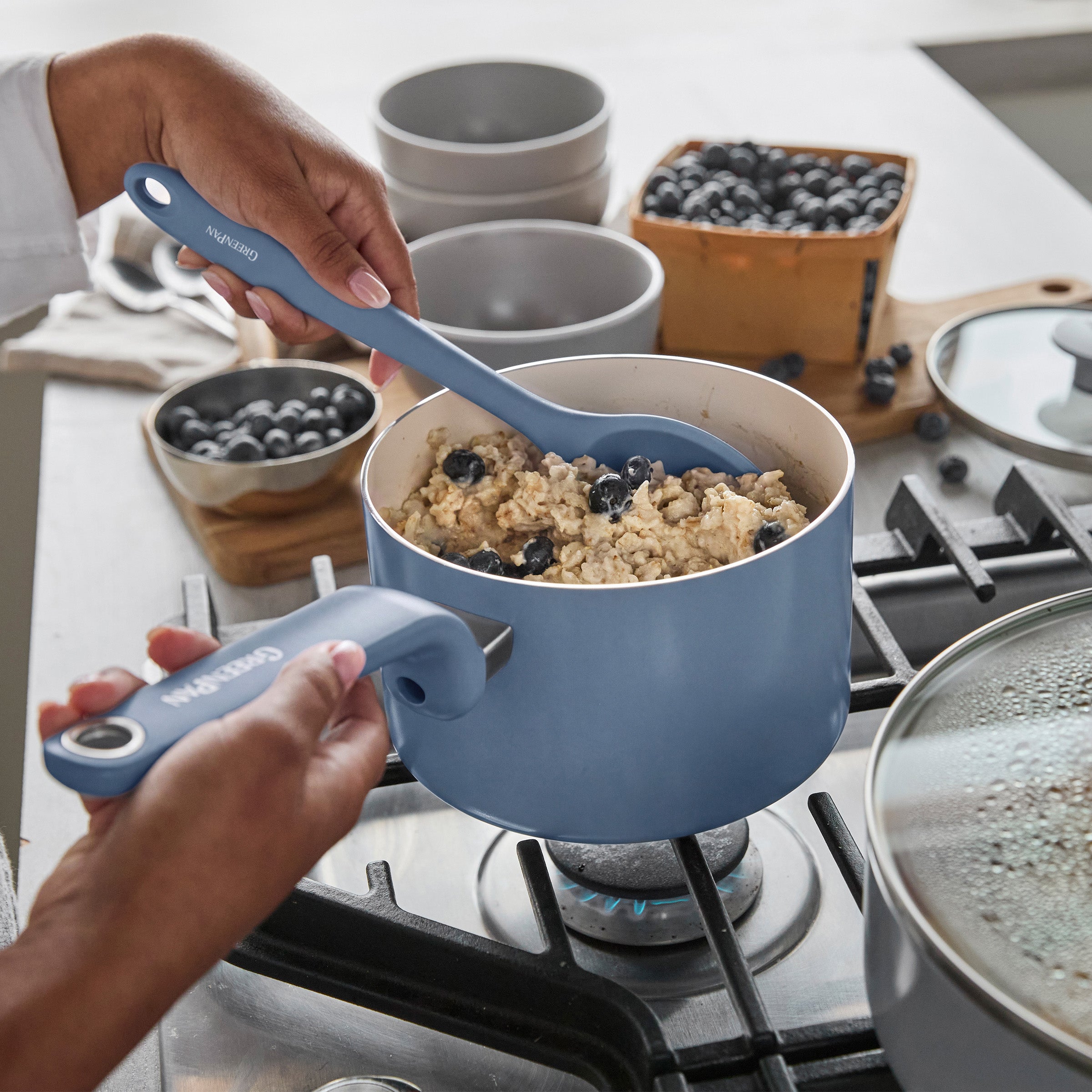 Oatmeal with blueberries being stirred in a blue ceramic pot; matching bowls and fresh berries are on the counter nearby.