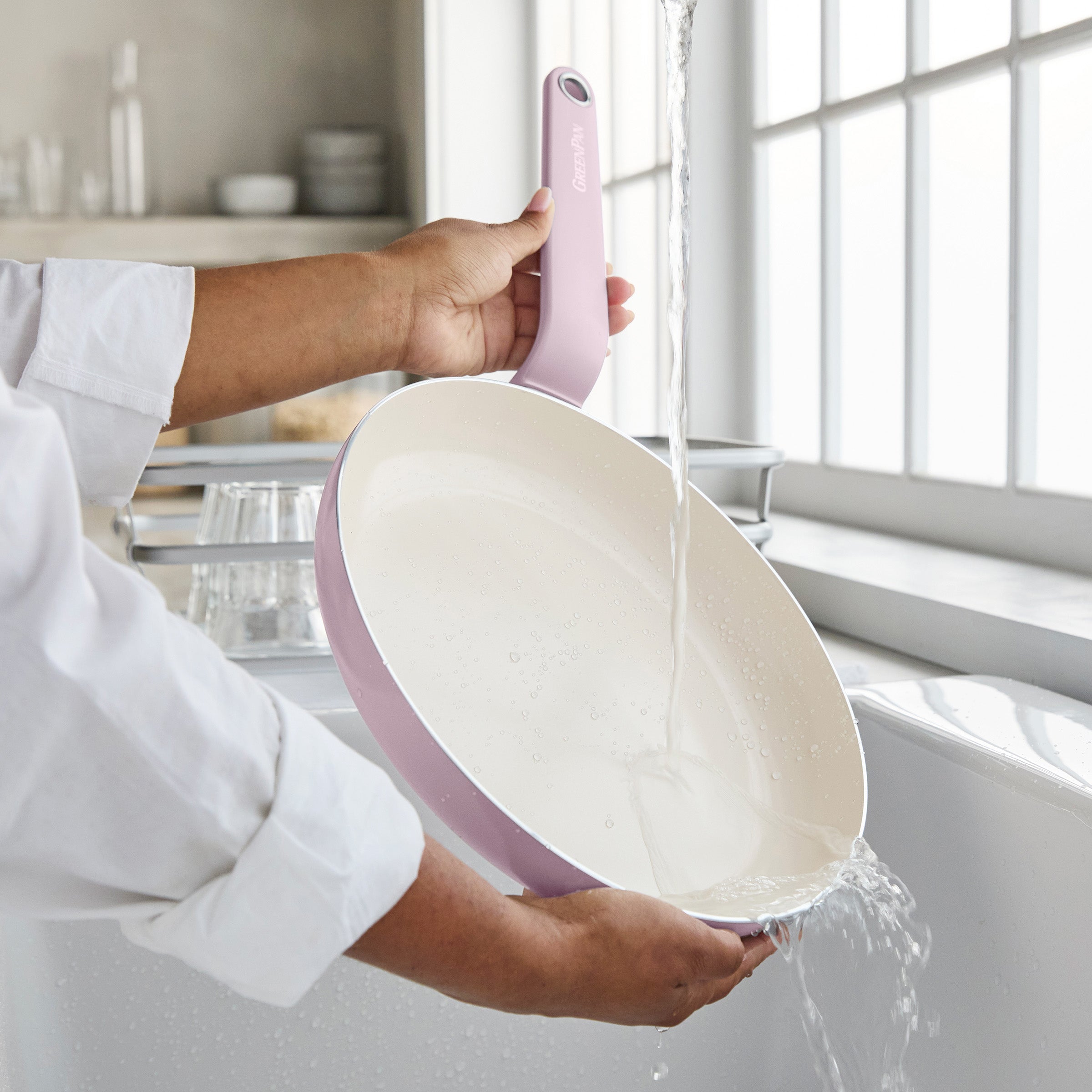 A pink 12-piece ceramic nonstick cookware set being rinsed in a bright kitchen with large windows.