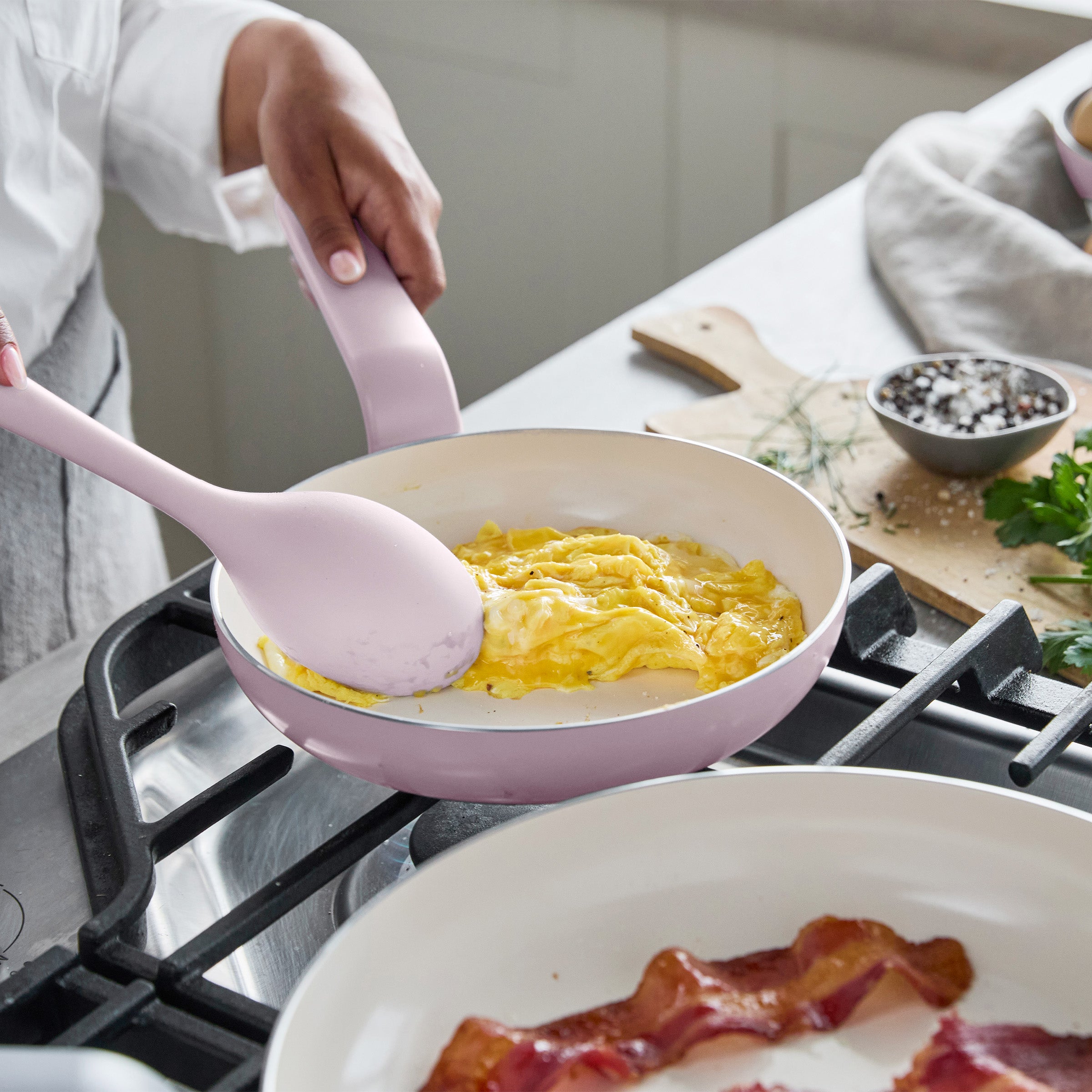 Scrambled eggs cooking in a pink ceramic nonstick pan, with strips of bacon beside it.