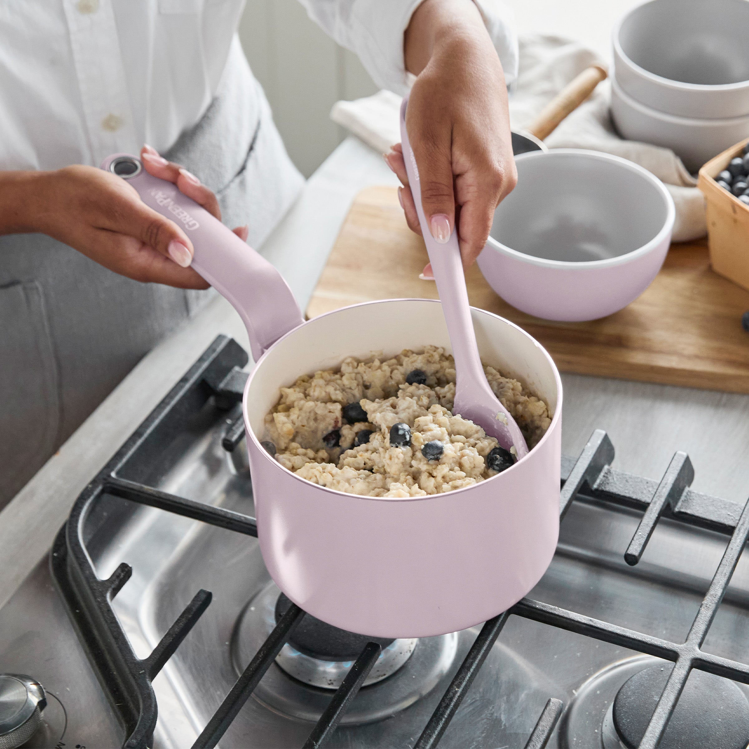 Pink ceramic nonstick pot with oatmeal and blueberries being stirred, bowls and fresh berries on the stove nearby.