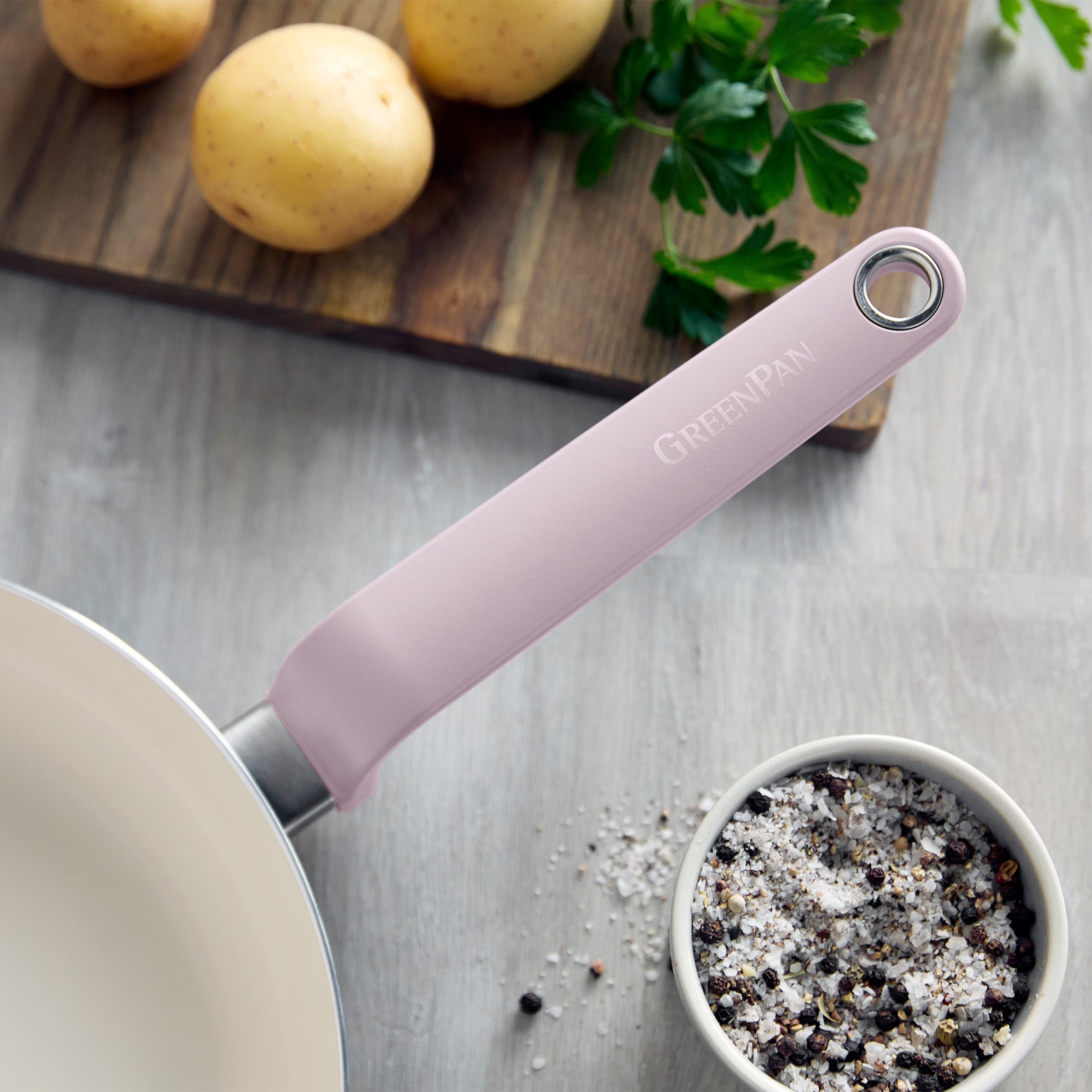 Pink 12-piece ceramic nonstick cookware set on a wooden surface beside potatoes, parsley, and pepper salt.