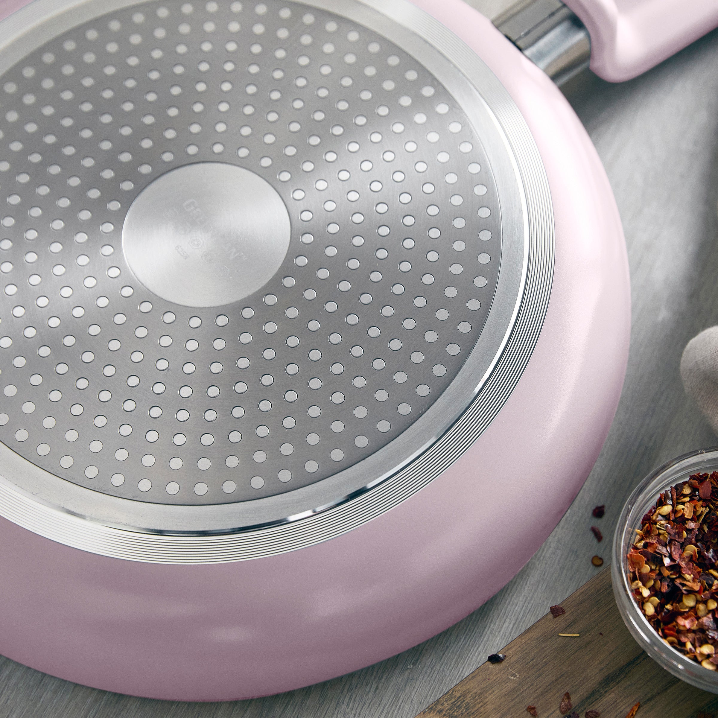 Pink 12-piece ceramic nonstick cookware set with dotted metal base, shown close up next to chili flakes.