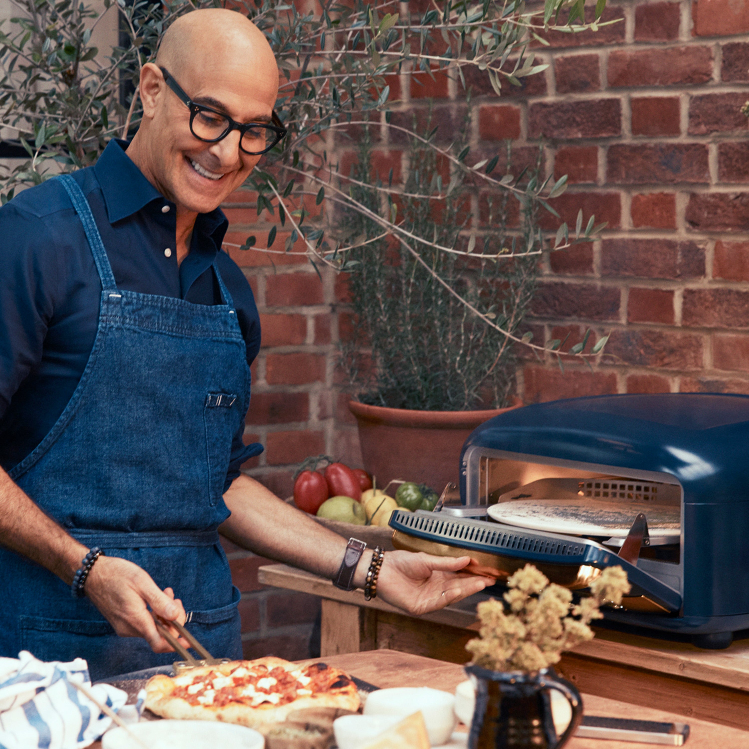 Stanley Tucci in a blue apron cooking pizza outdoors with a brick wall and plants in the background.