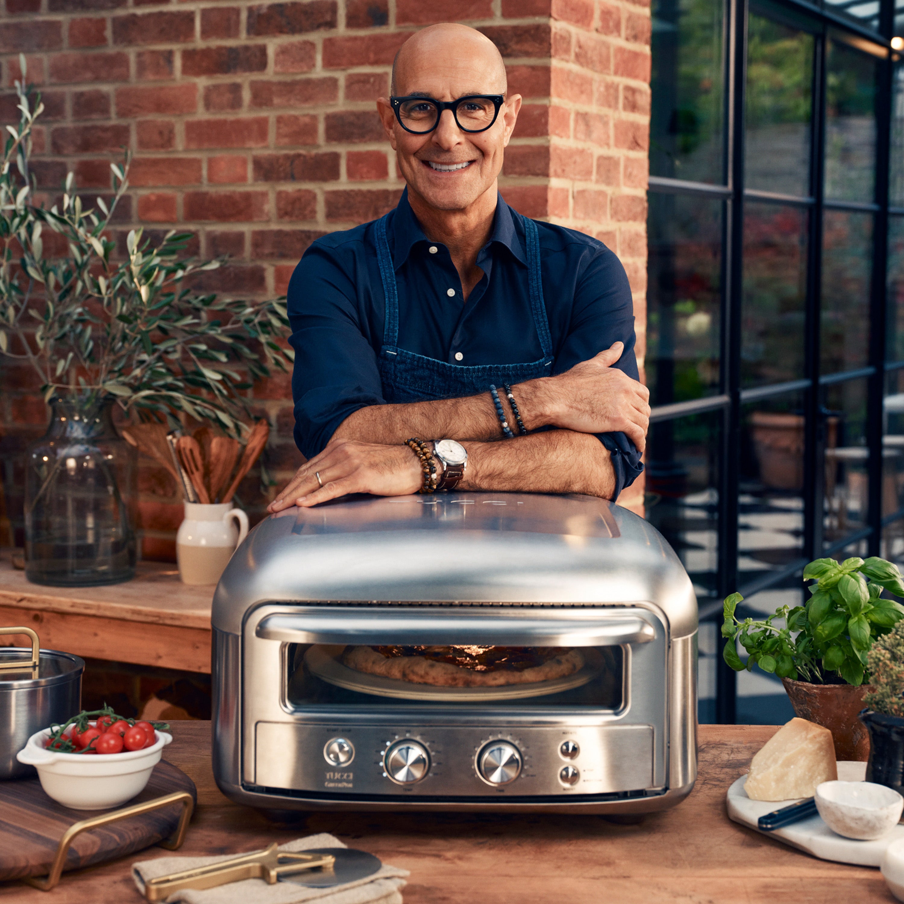 Stanley Tucci standing behind a stainless steel pizza oven in an outdoor setting