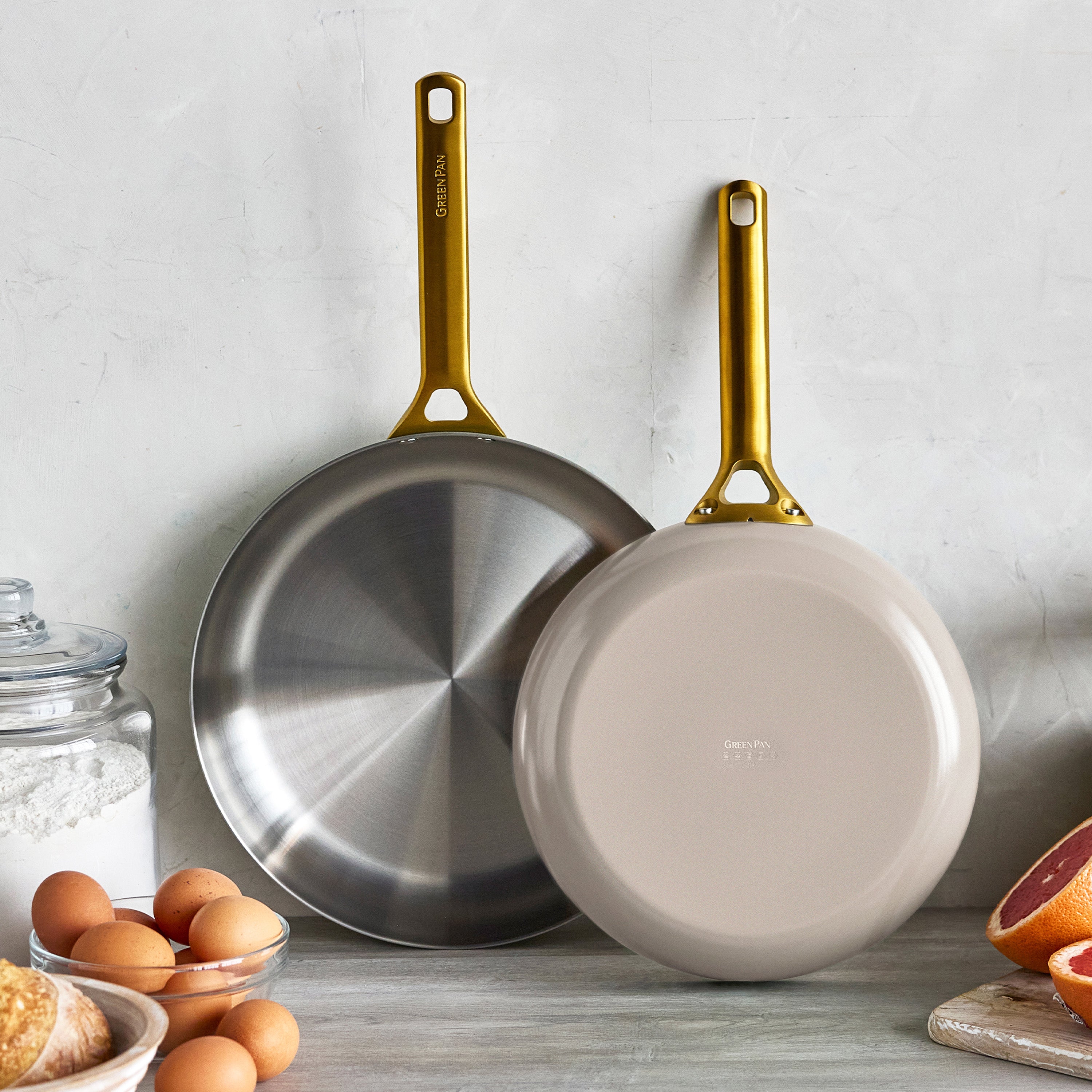 Set of two uncoated stainless steel frypans in macadamia with gold handles hanging above a kitchen counter with ingredients.