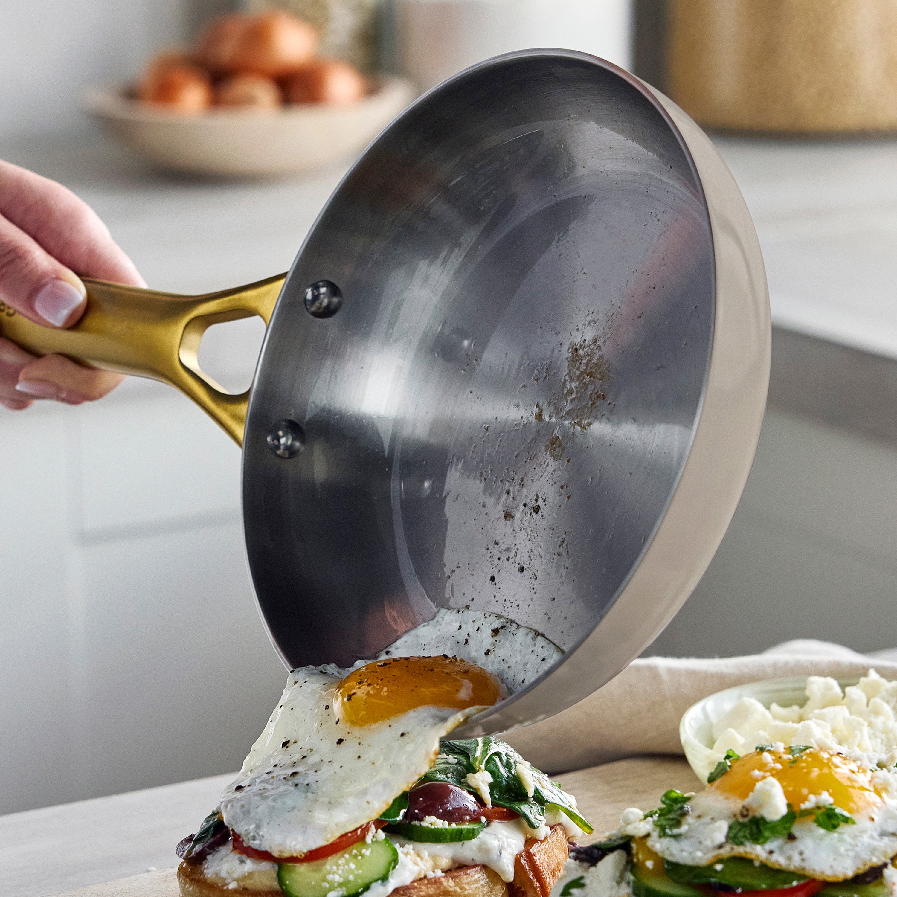 A hand slides a fried egg from a stainless steel frypan onto toast topped with vegetables; both 10″ and 12″ pans are shown.