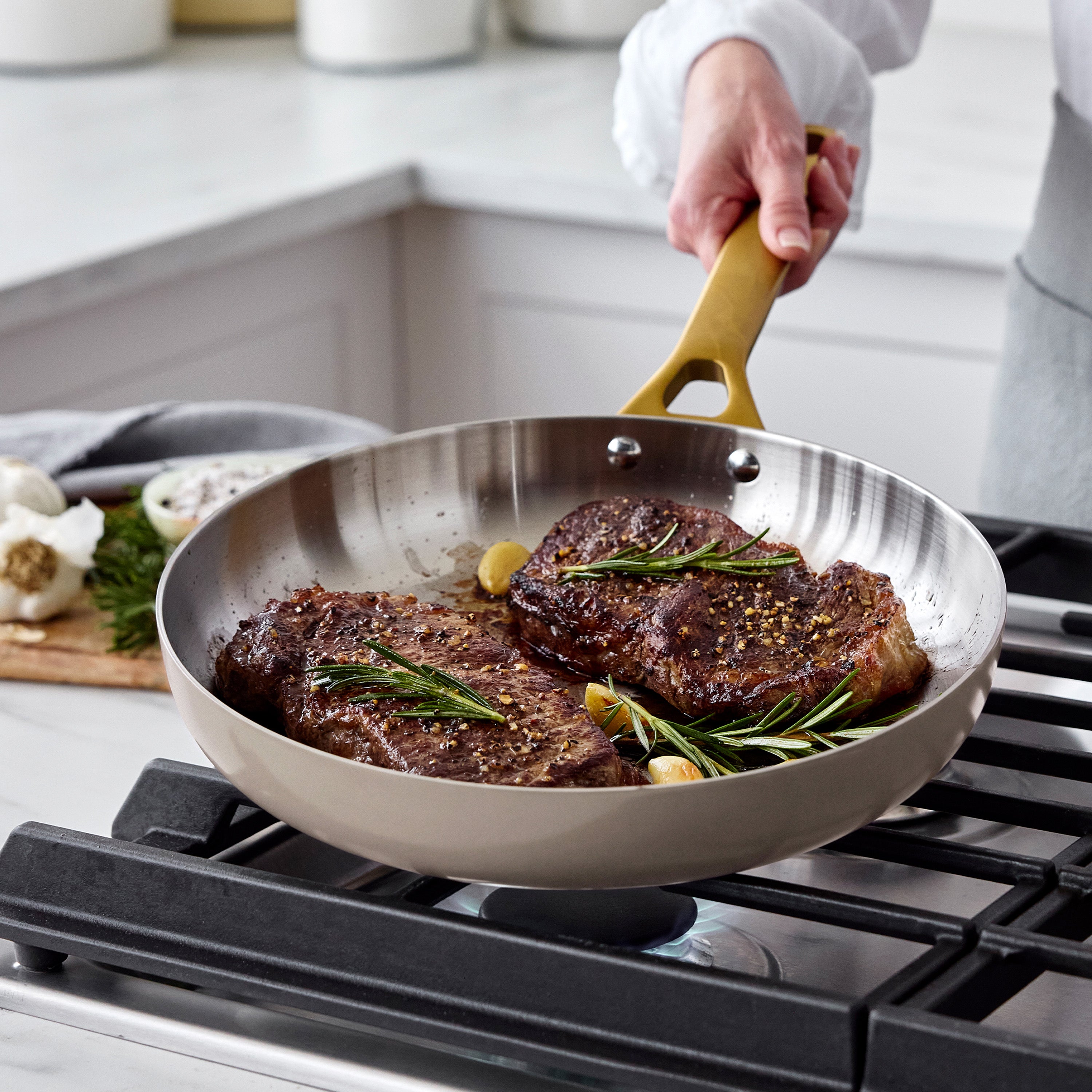 Hand holds two uncoated stainless steel frypans with seared steaks, garlic, and rosemary; Macadamia handles visible.