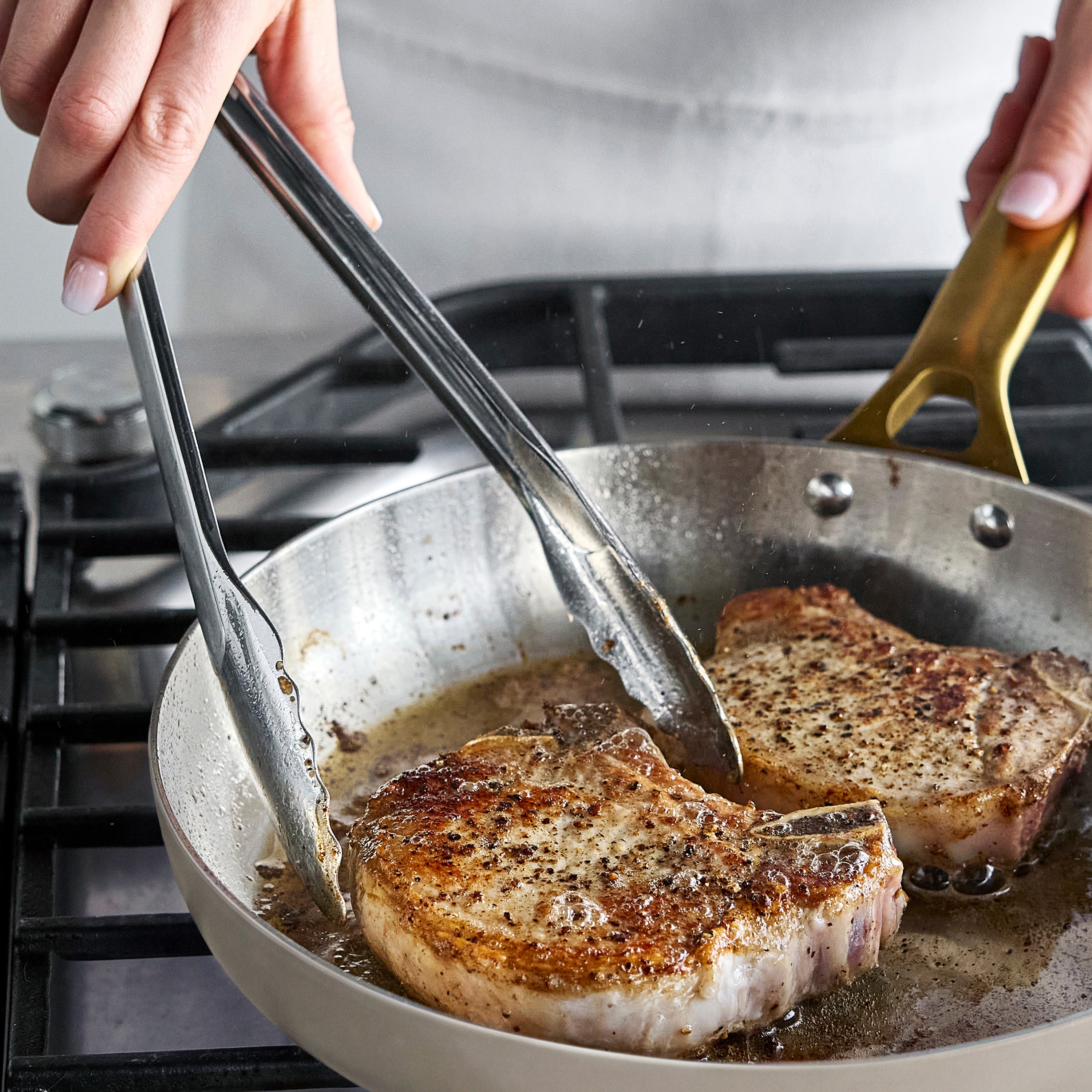 Two pork chops cooking in a stainless steel frypan set on a stovetop, held by tongs.