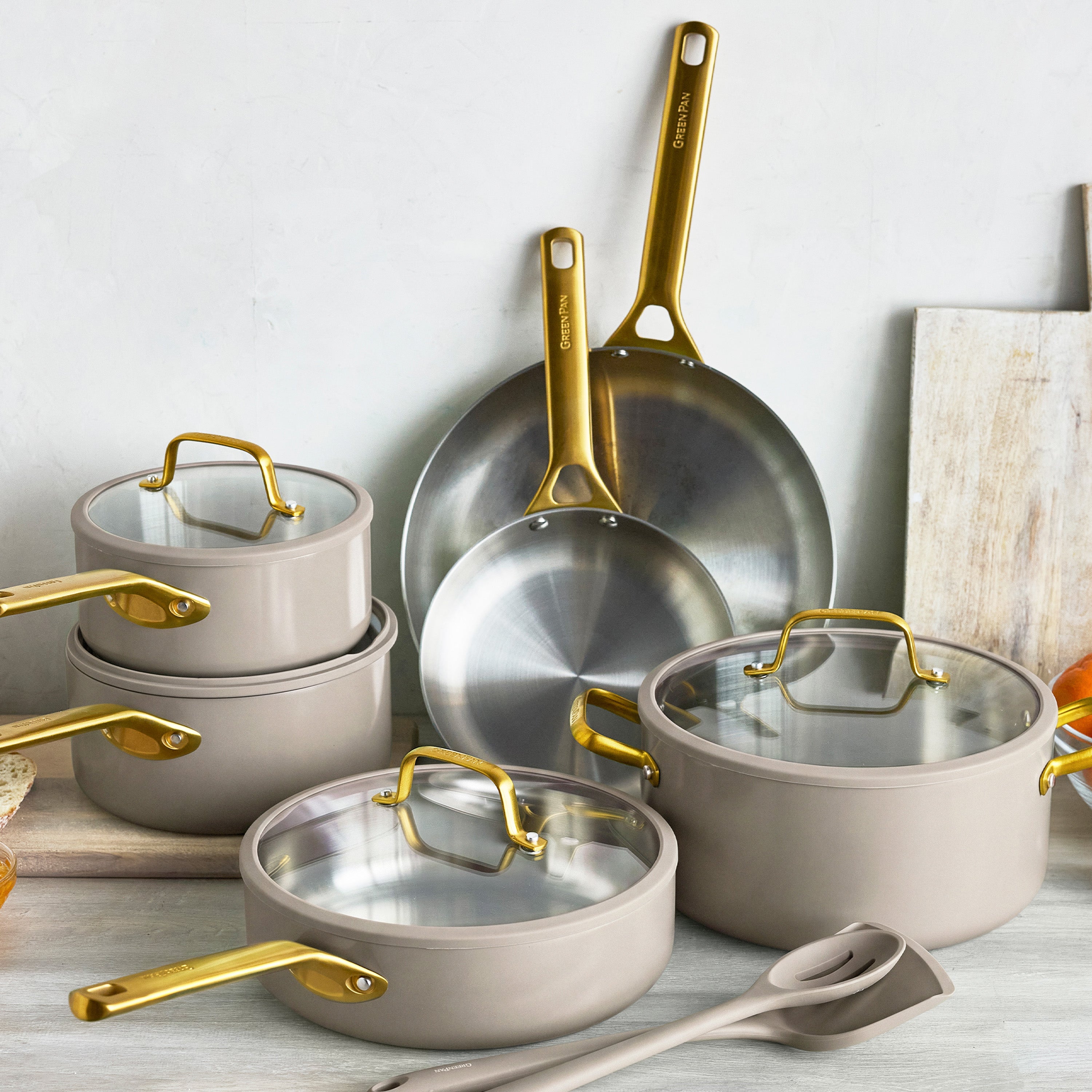 12-piece stainless steel cookware set in macadamia with gold handles and glass lids arranged on a kitchen counter.