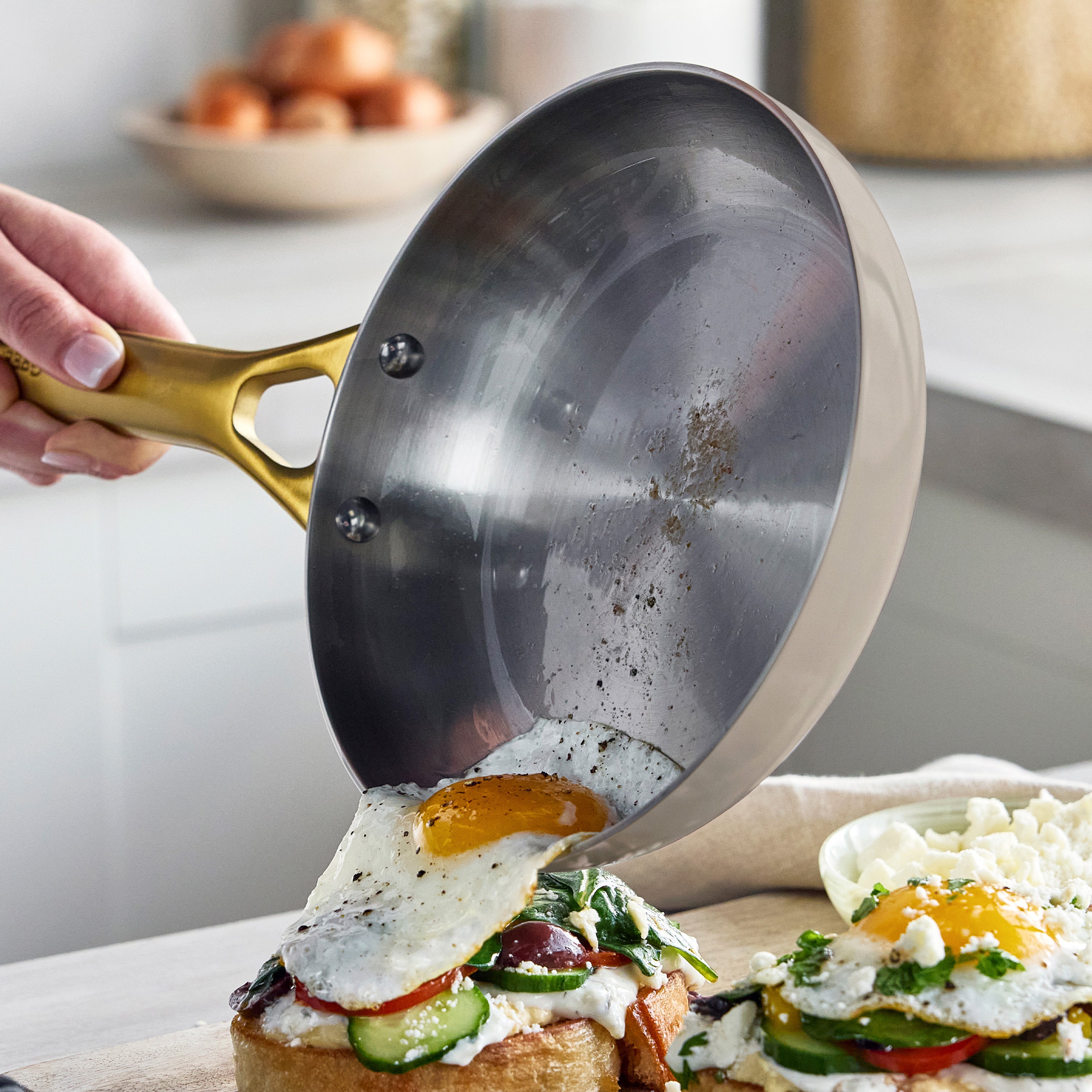 Hand using stainless steel frying pan to serve a fried egg onto veggie toast; part of 12-piece cookware set, Macadamia finish visible.