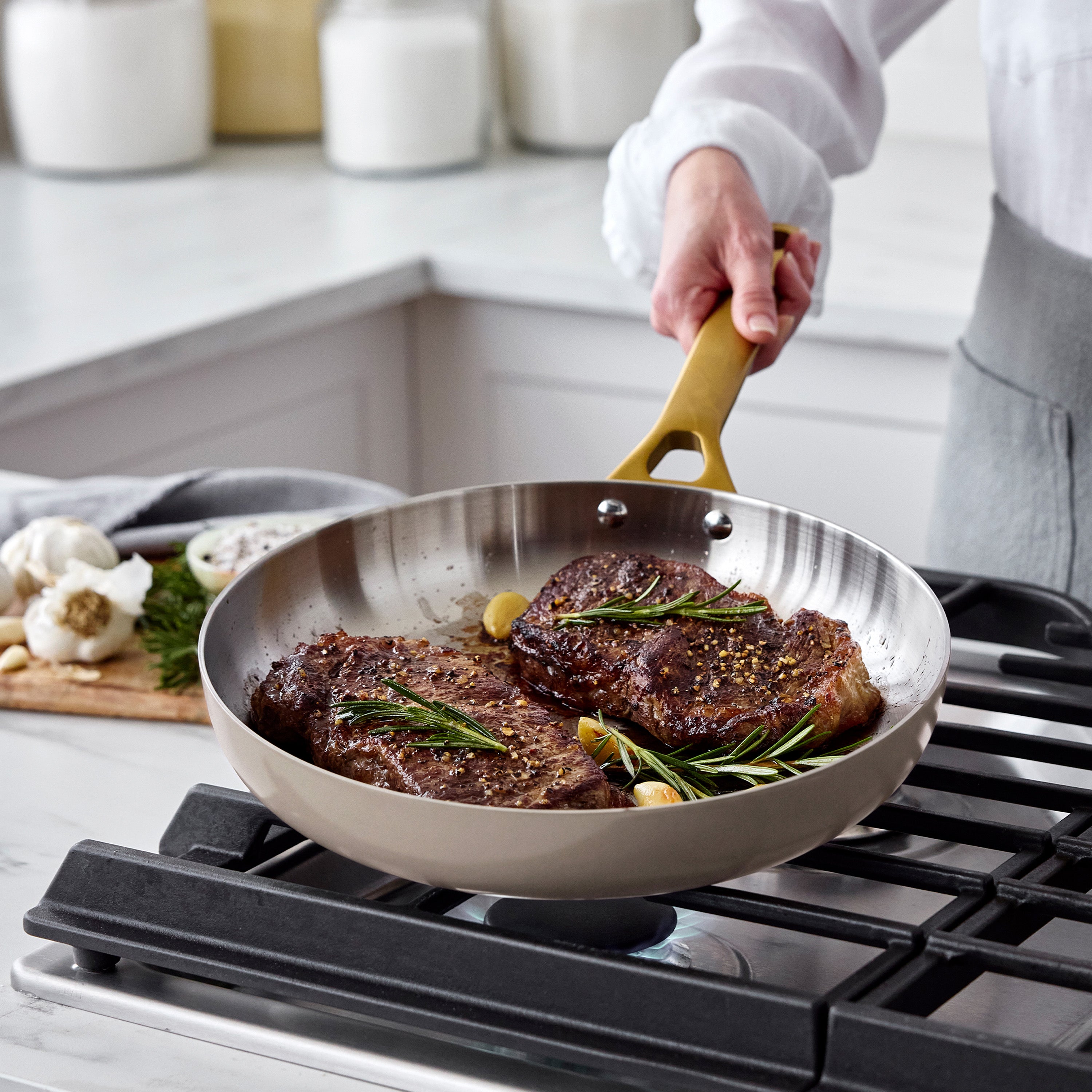 Two steaks with rosemary cook in a stainless steel pan from a 12-piece set; macadamia nuts and garlic beside the cookware.