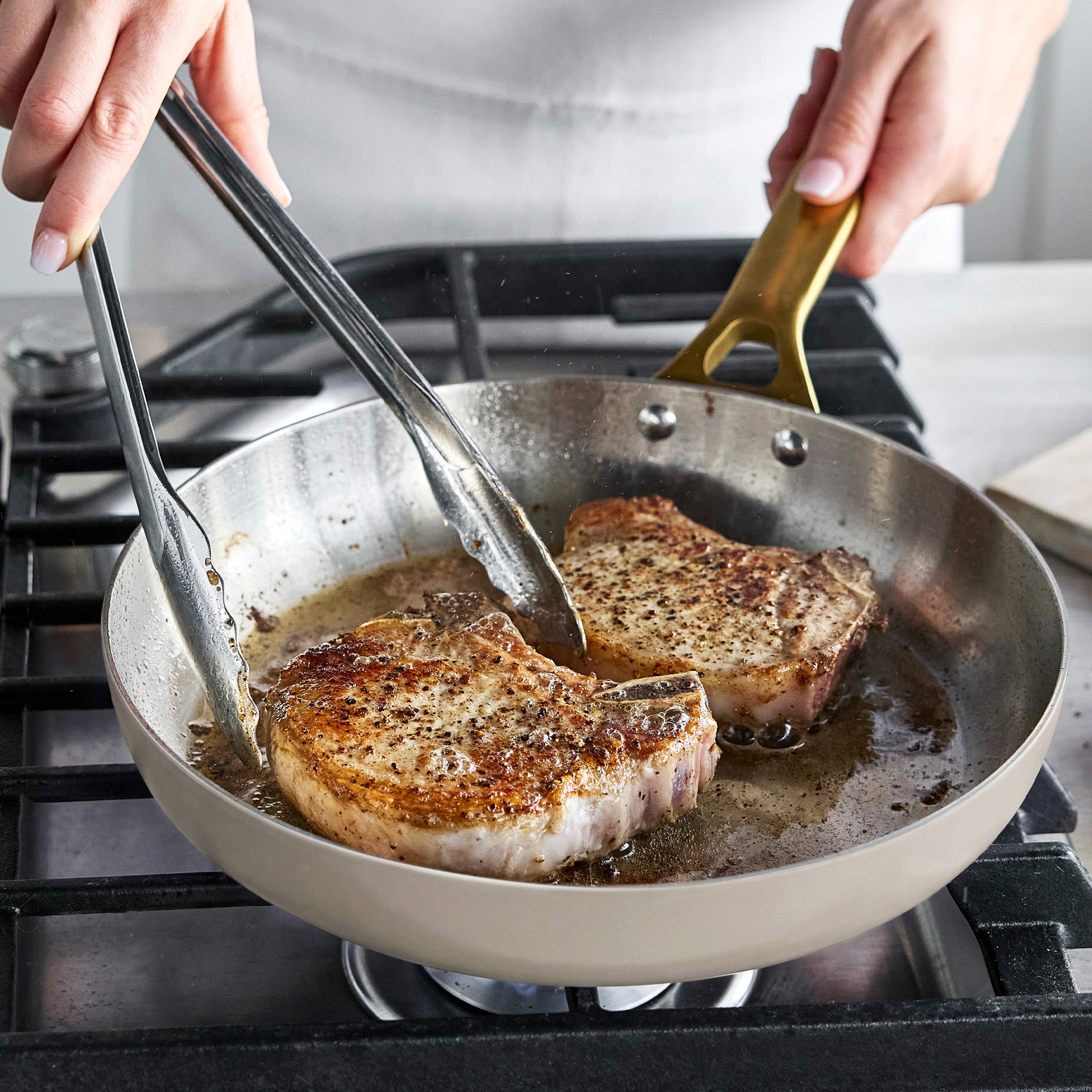 Pork chops being seared with tongs in a stainless steel pan from the 12-piece Macadamia cookware set.