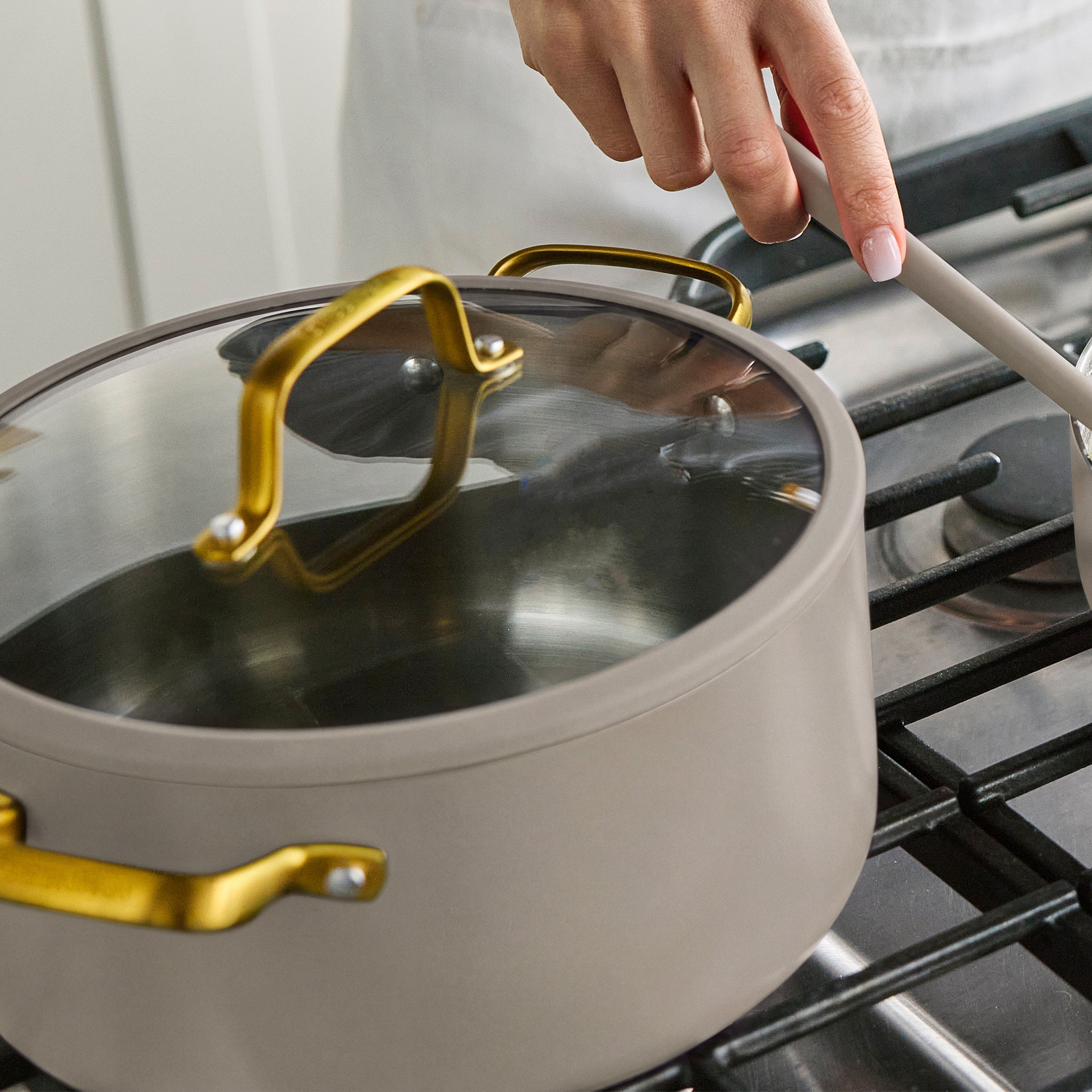 A hand stirs food on a stove beside a 12-piece stainless steel cookware set in macadamia color with gold handles.