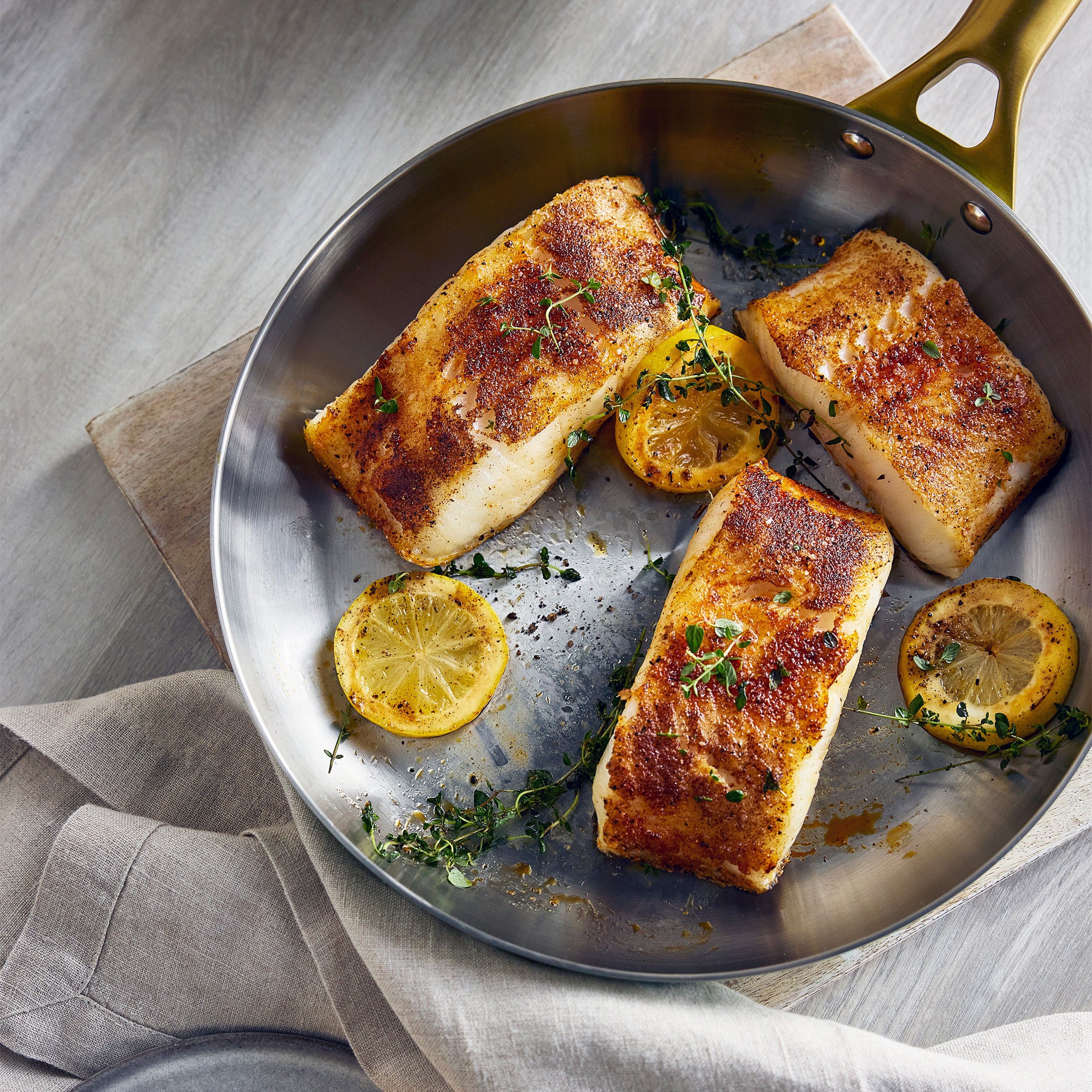 Three cooked fish fillets with lemon and herbs in a stainless steel pan on a wooden surface.