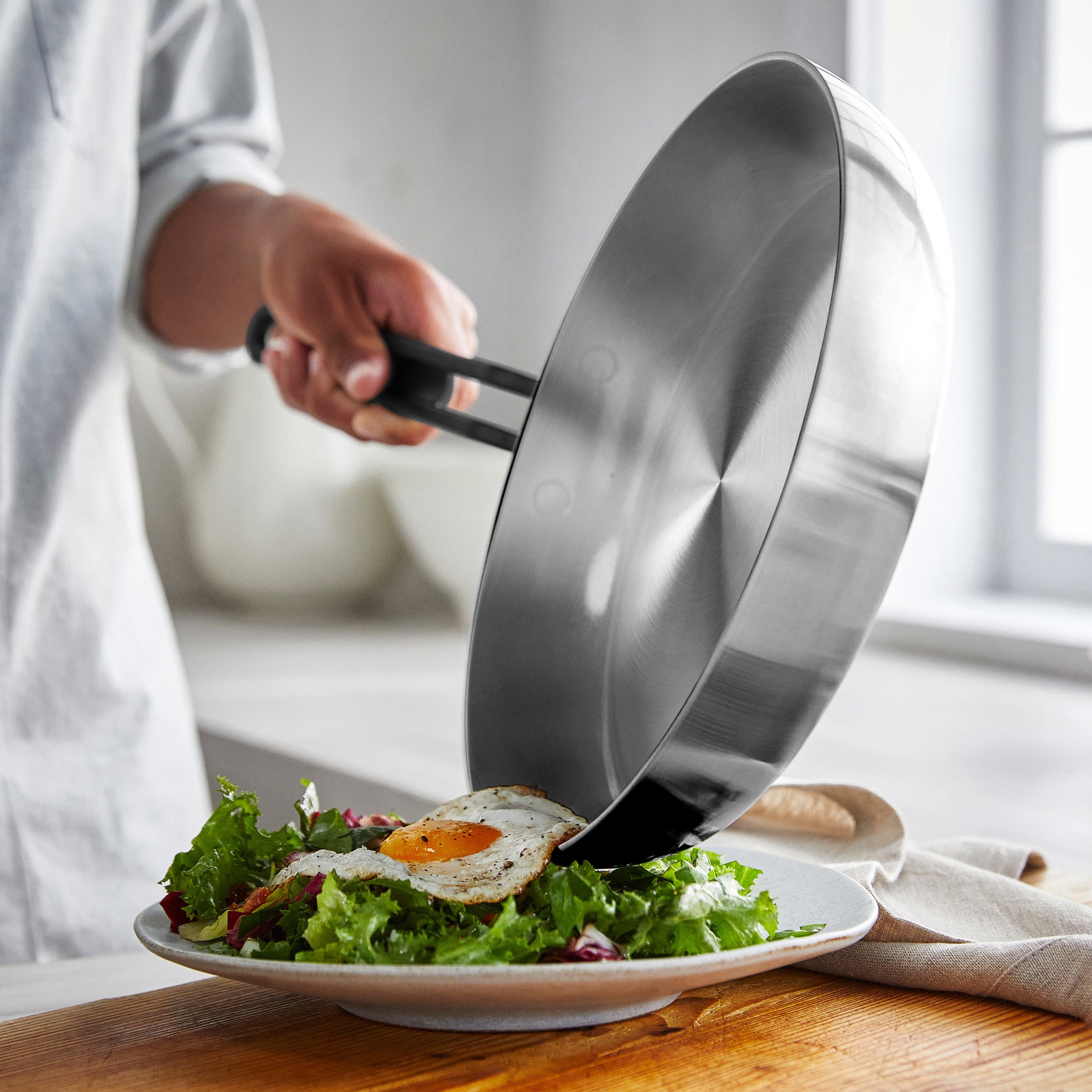 A person slides a fried egg from a white ceramic nonstick pan onto a green salad.