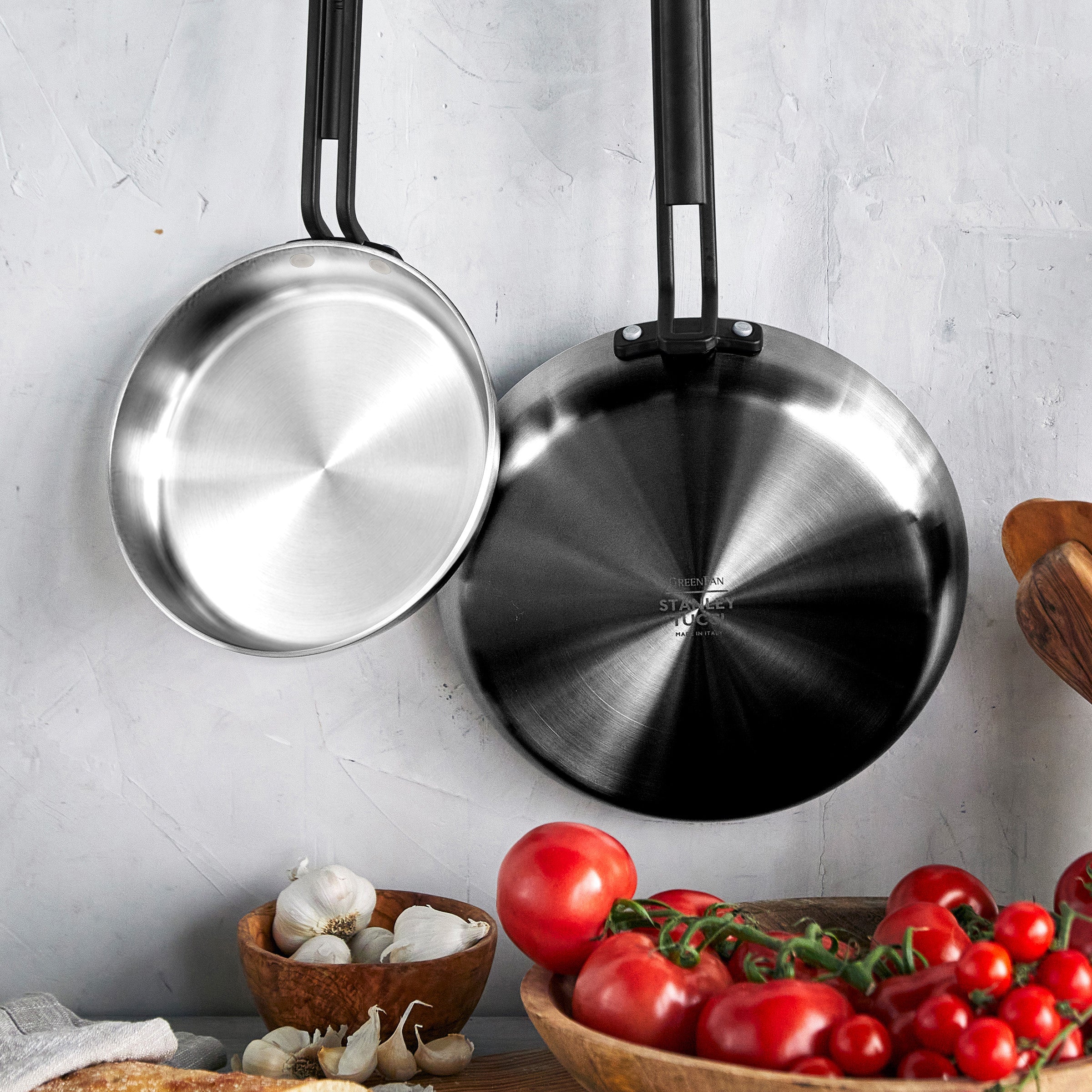 Two Stanley Tucci pans hanging above a table with tomatoes, garlic, mushrooms, and a wooden bowl.