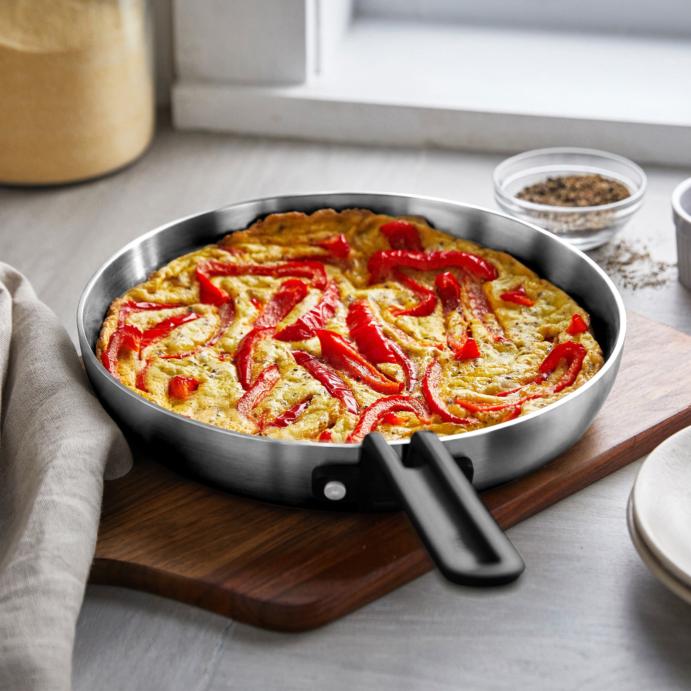 Red pepper frittata in a Stanley Tucci skillet on a board near spices and a window.